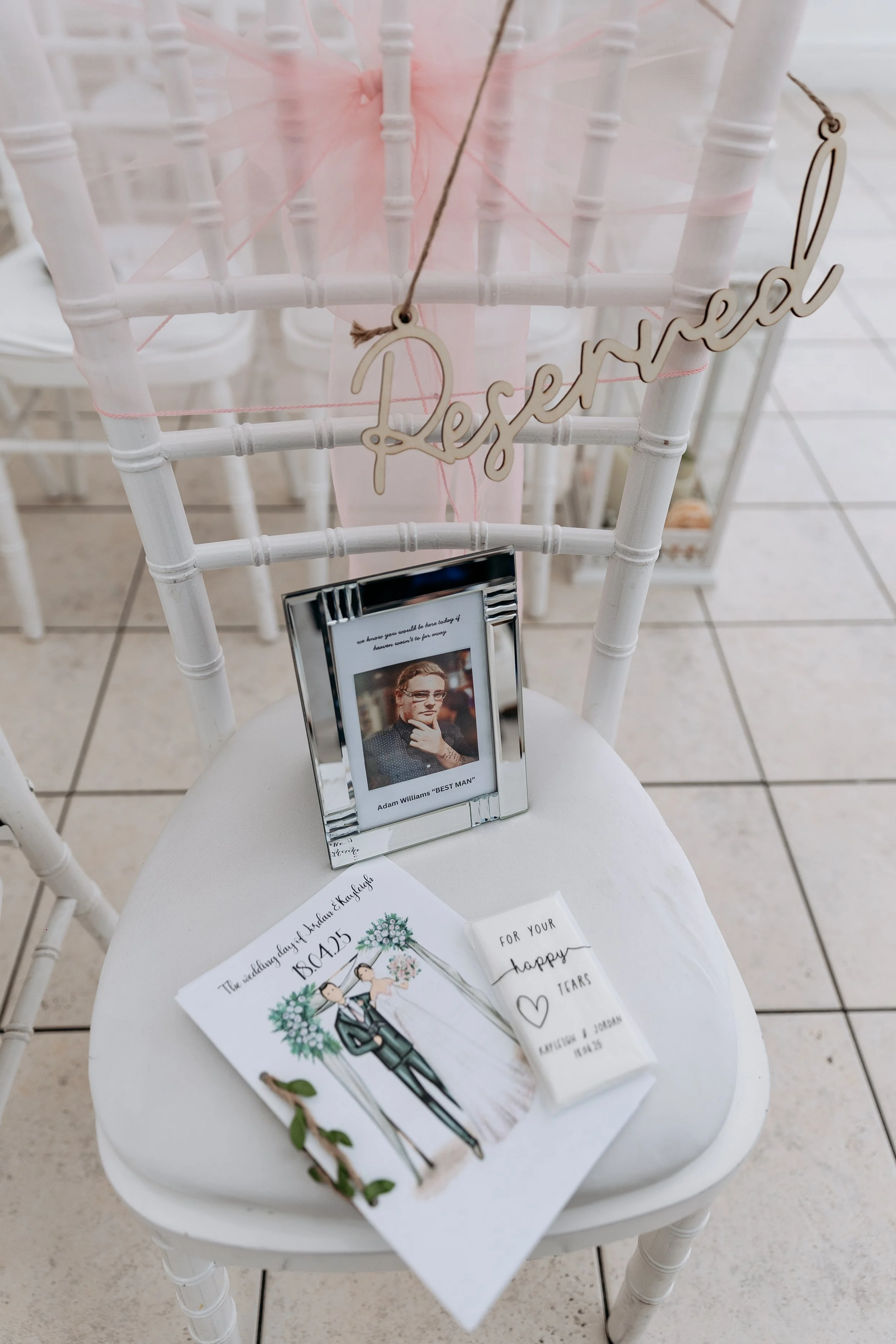 A white chair decorated for a wedding with a pink tulle bow, a wooden 'Reserved' sign hanging, a framed photograph of a man, a wedding program, and a small box with an inscription.