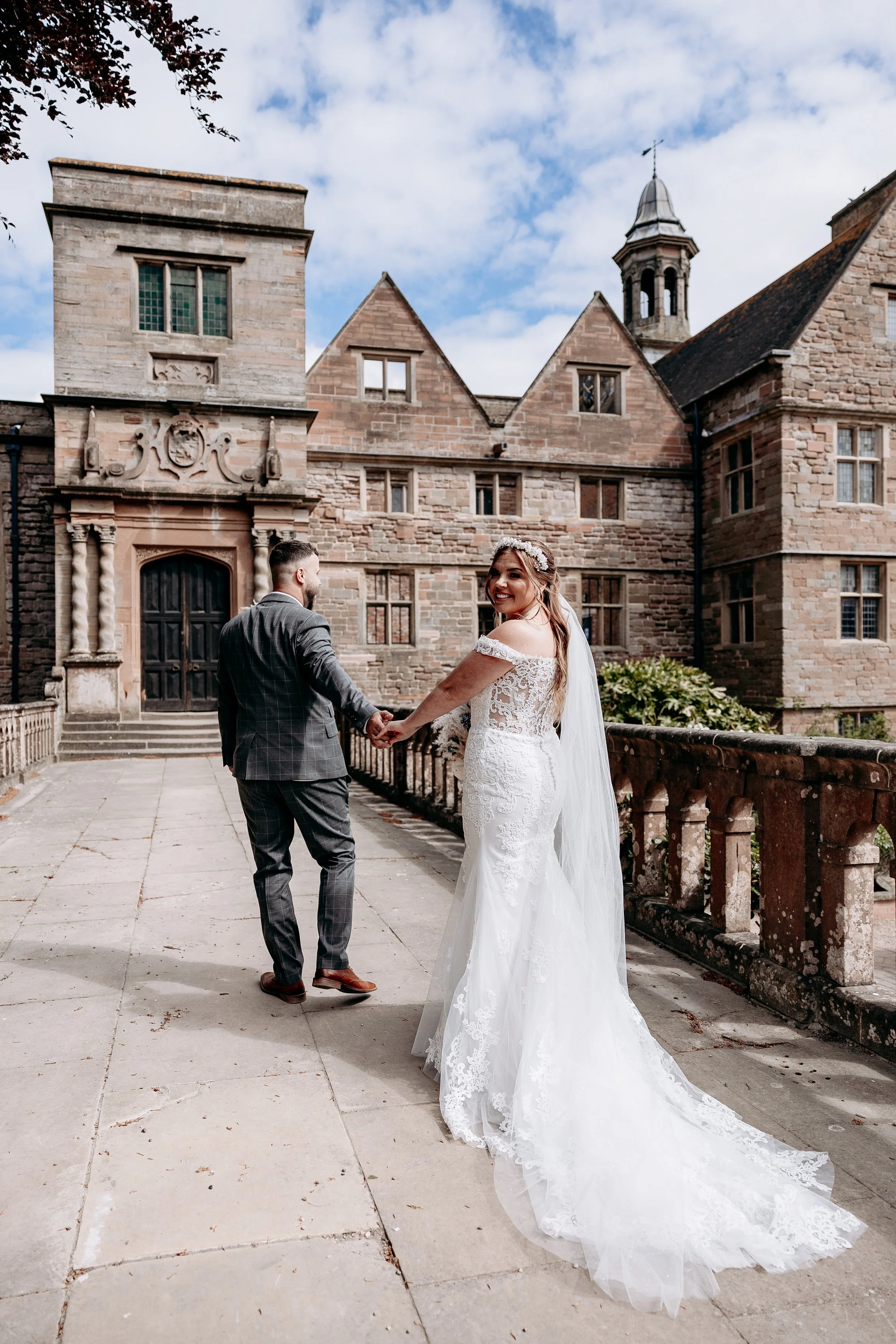 A bride and groom holding hands, standing on a stone walkway outside a historic stone castle or mansion, under a partly cloudy sky.