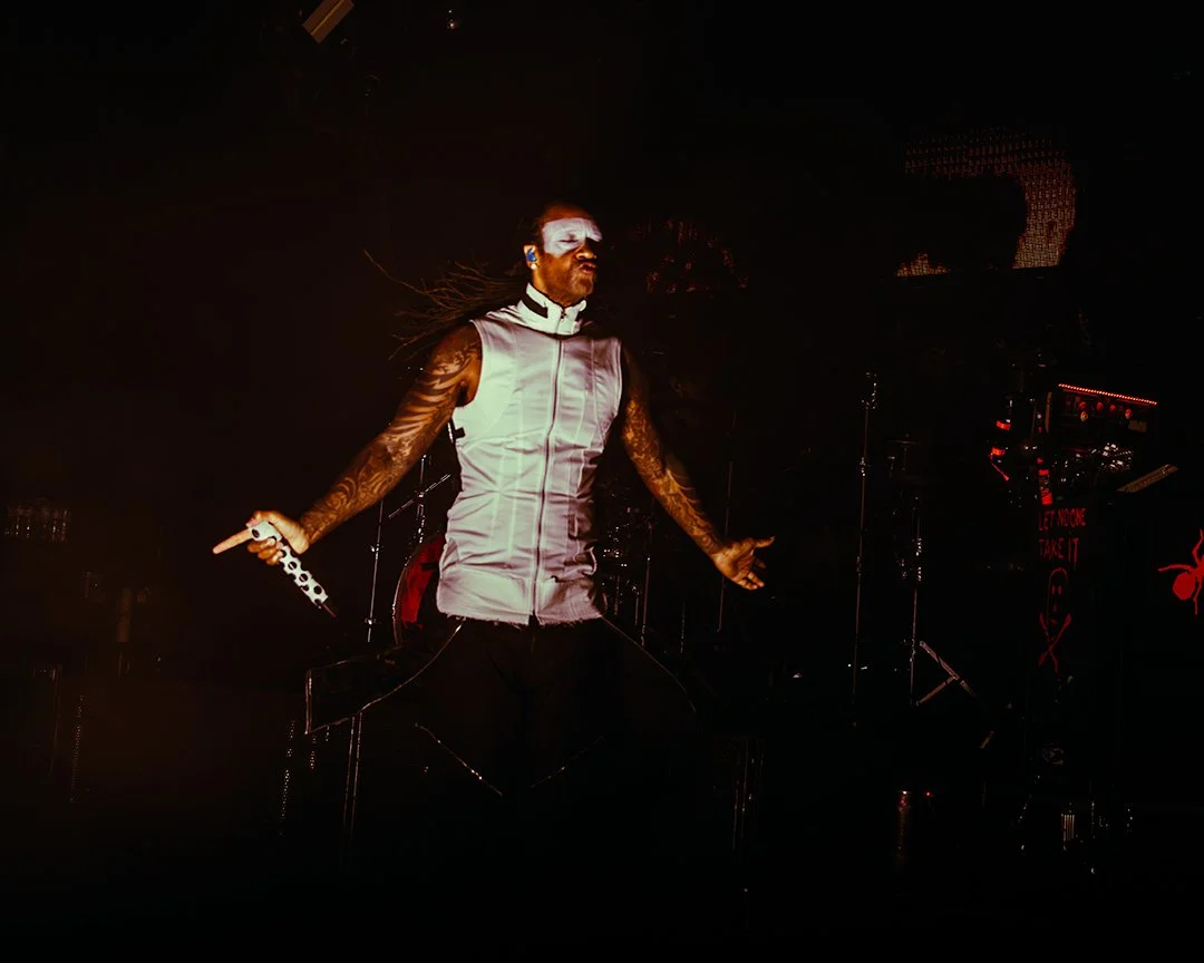 A male performer with face paint and tattoos holding a microphone on stage during a concert, with dark background and stage lights.