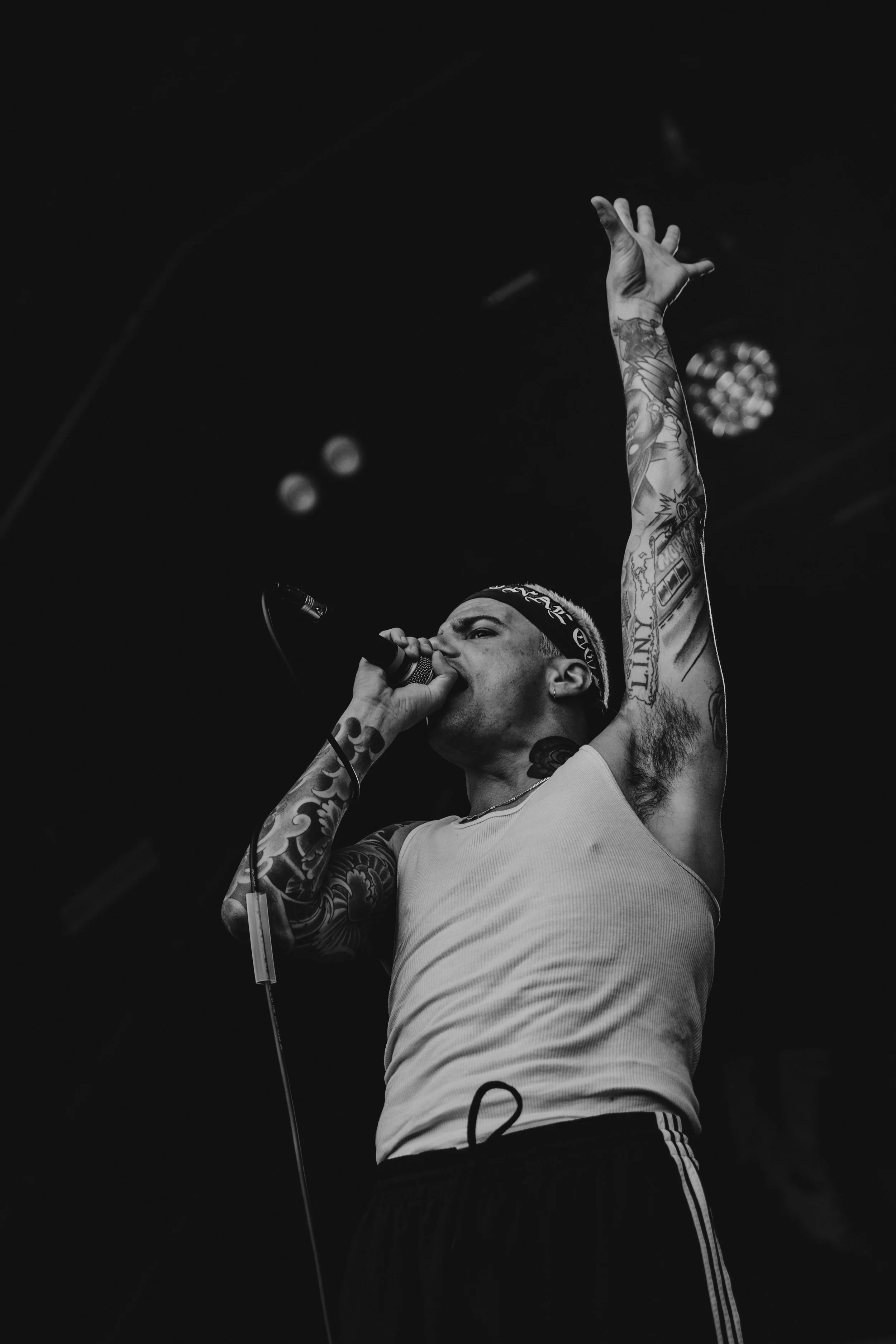 A male singer performs on stage, holding a microphone close to his mouth, with one arm raised in the air, wearing a sleeveless shirt, a bandana, and displaying tattoos on his arms.