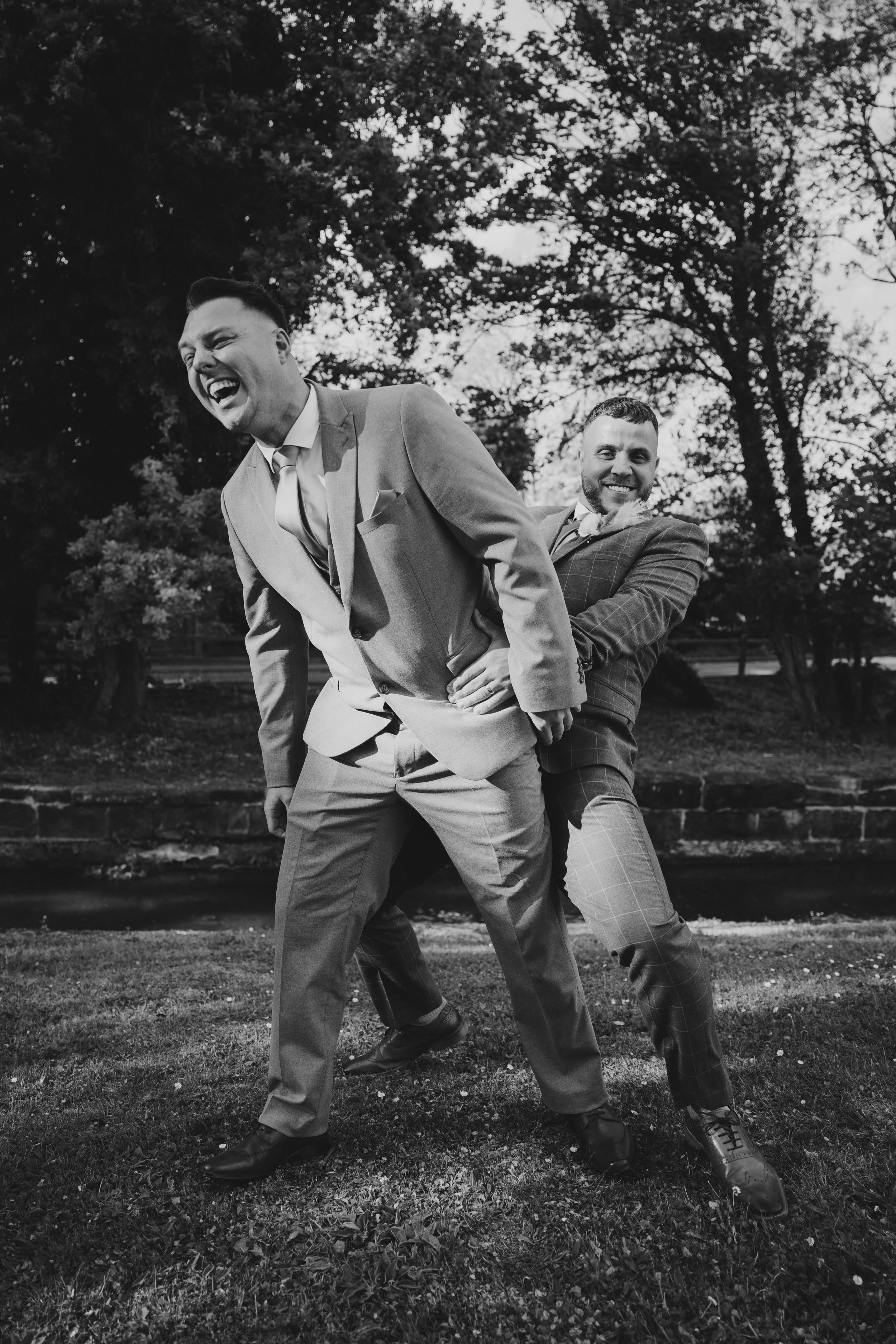 Two men in suits laughing and playfully posing outdoors on grass near a small water body and trees, black and white photo.