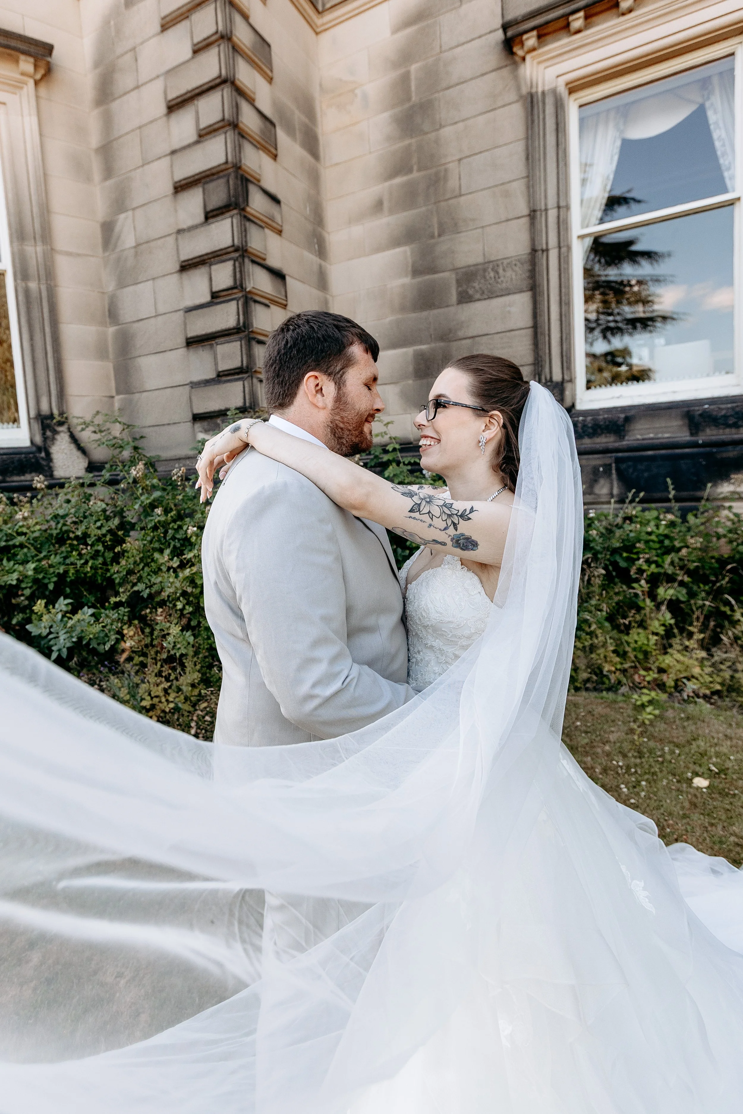 Bride and groom kiss outdoors in wedding attire, with the bride wearing a white gown and veil, and the groom in a light-colored suit, in front of an old stone building with windows and greenery.