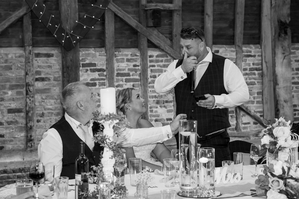 Black and white photo of a wedding reception with three people sitting at a decorated table, one man standing and speaking into a microphone, another woman reaching out to him, and a man looking on, in a rustic venue with brick walls and string light
