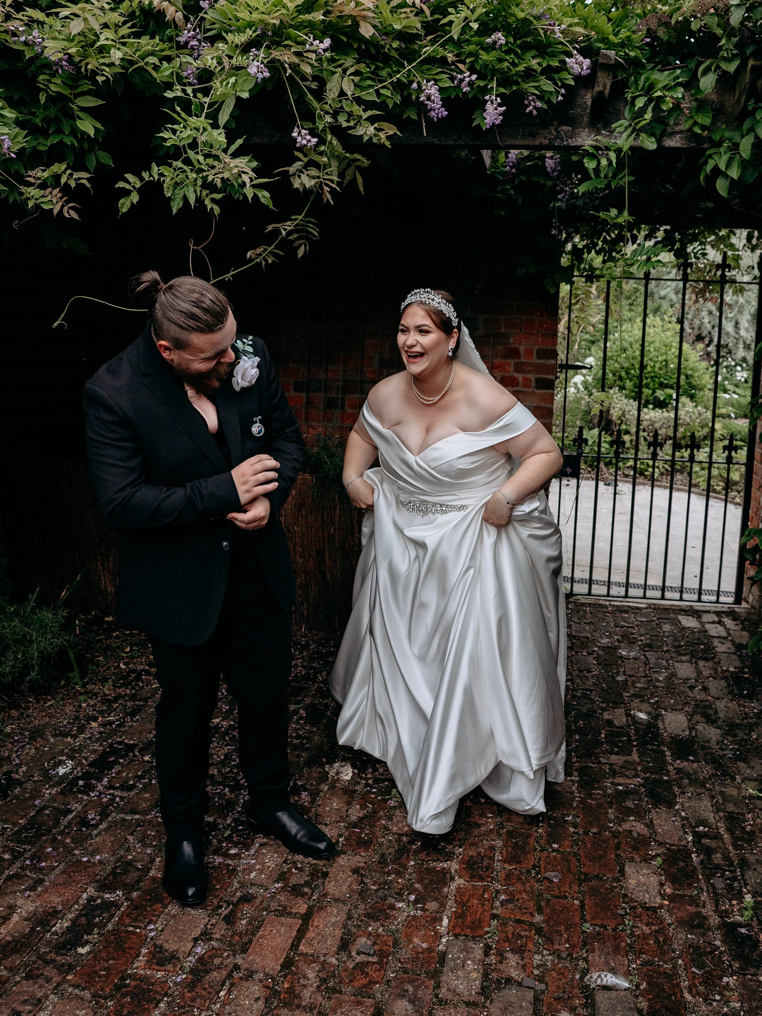 Bride in white wedding gown and tiara laughing with groom in black suit outside in a garden area with greenery and brick ground.