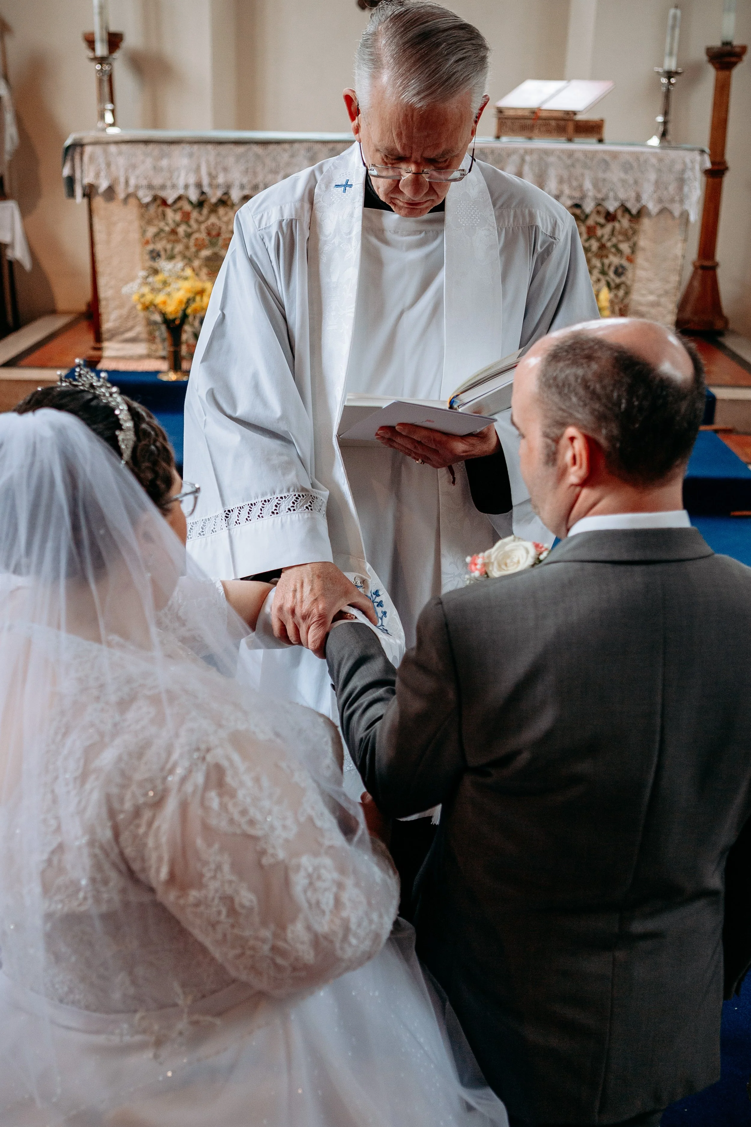 A priest conducts a wedding ceremony in a church, with a bride and groom holding hands and kneeling.