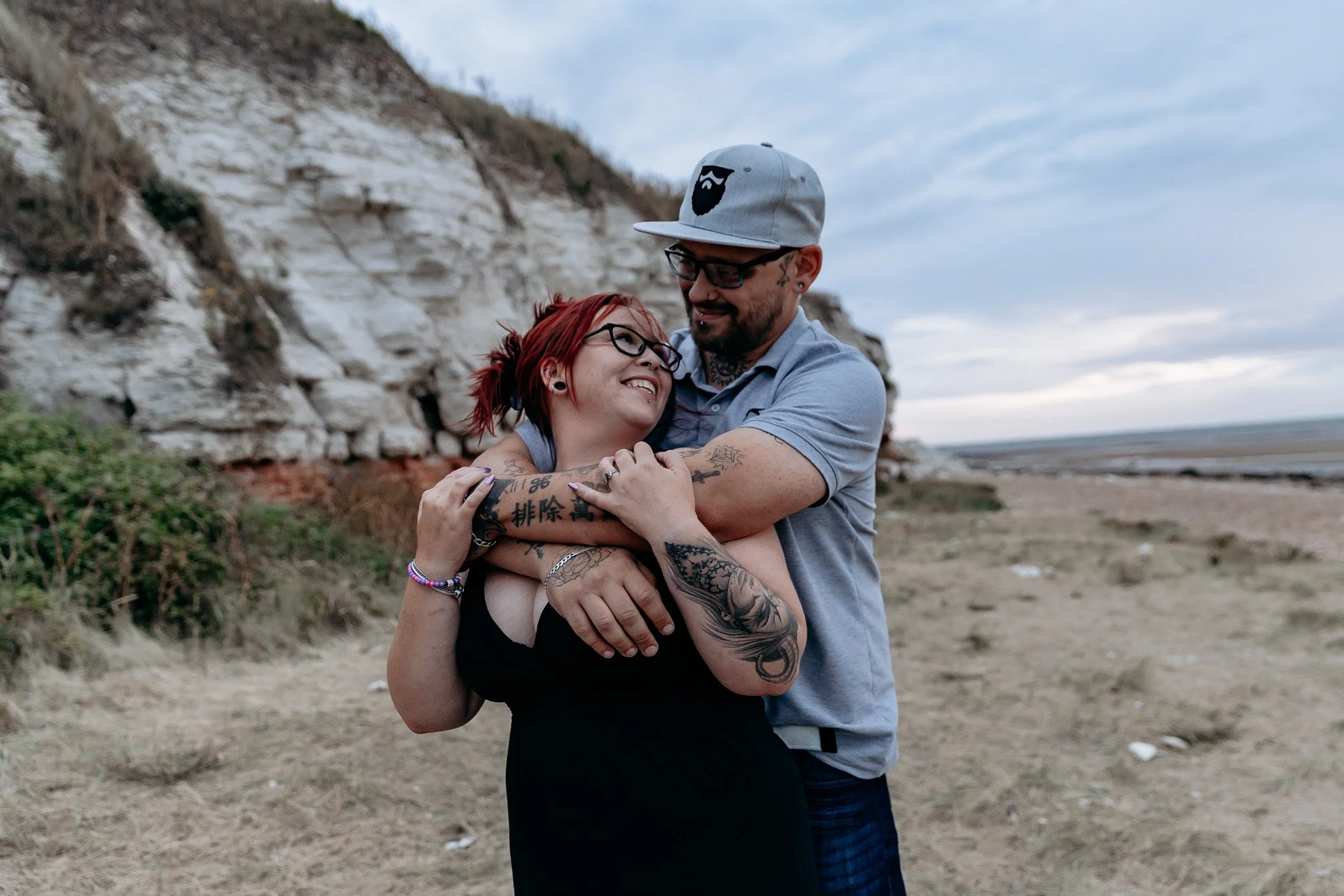 A smiling woman with red hair and glasses hugging a man with a beard, tattoos, and glasses at a beach with cliffs in the background.