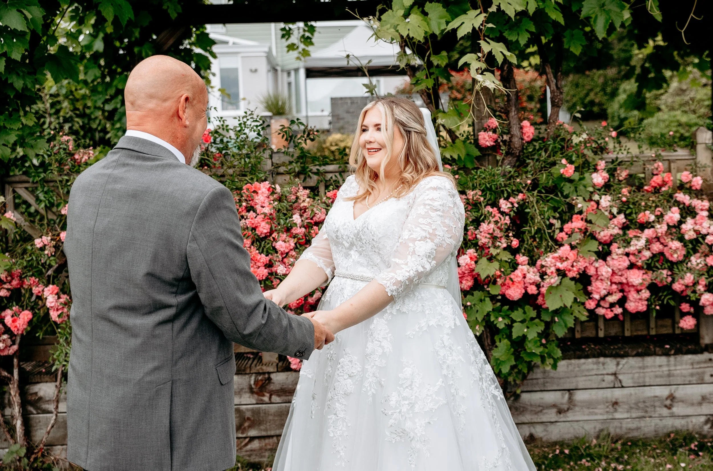 Bride in white lace gown holding hands with a man in a gray suit, standing in a garden with pink flowers.