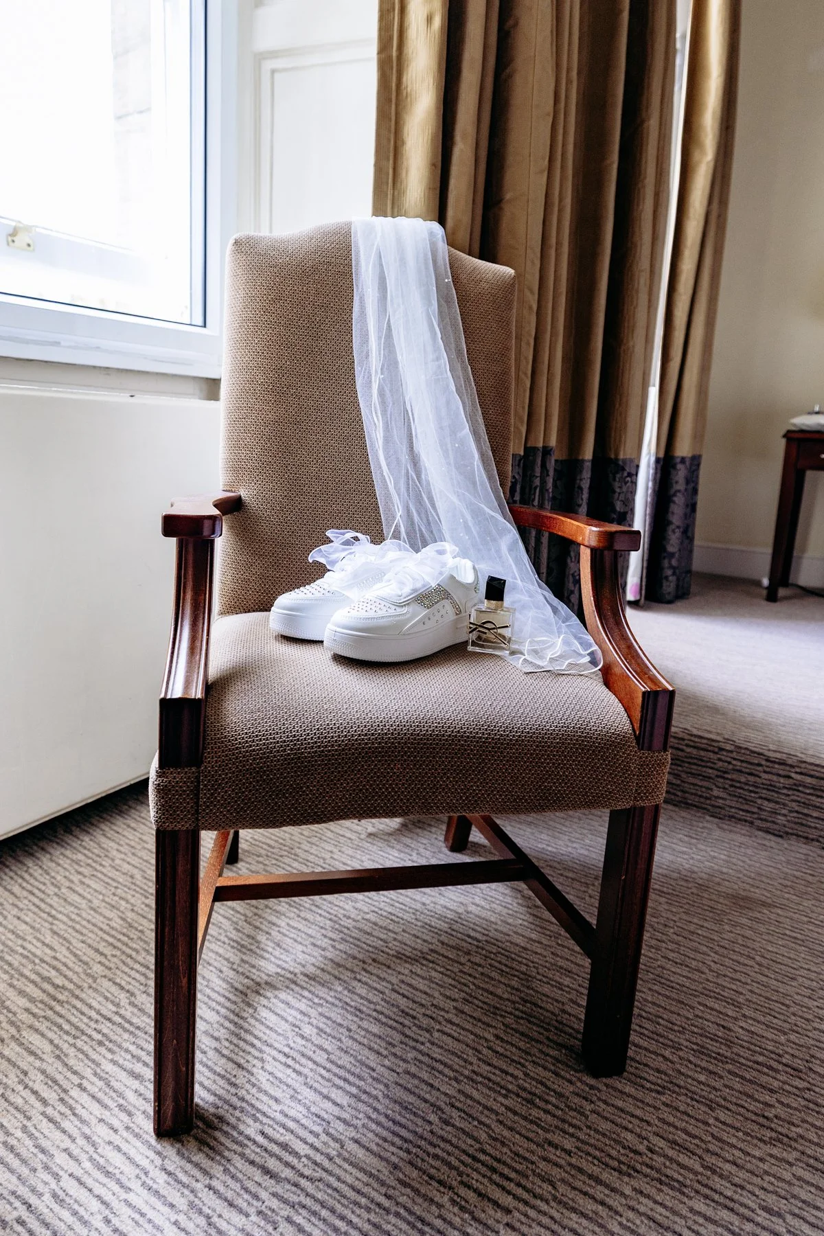 A beige armchair near a window with a veil, a pair of white sneakers, and a small bottle on the seat, indicating wedding preparations.