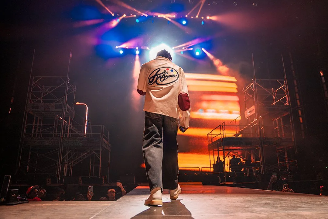 Person walking on stage with bright lights and large screen displaying colorful visuals in the background, during a concert or performance.