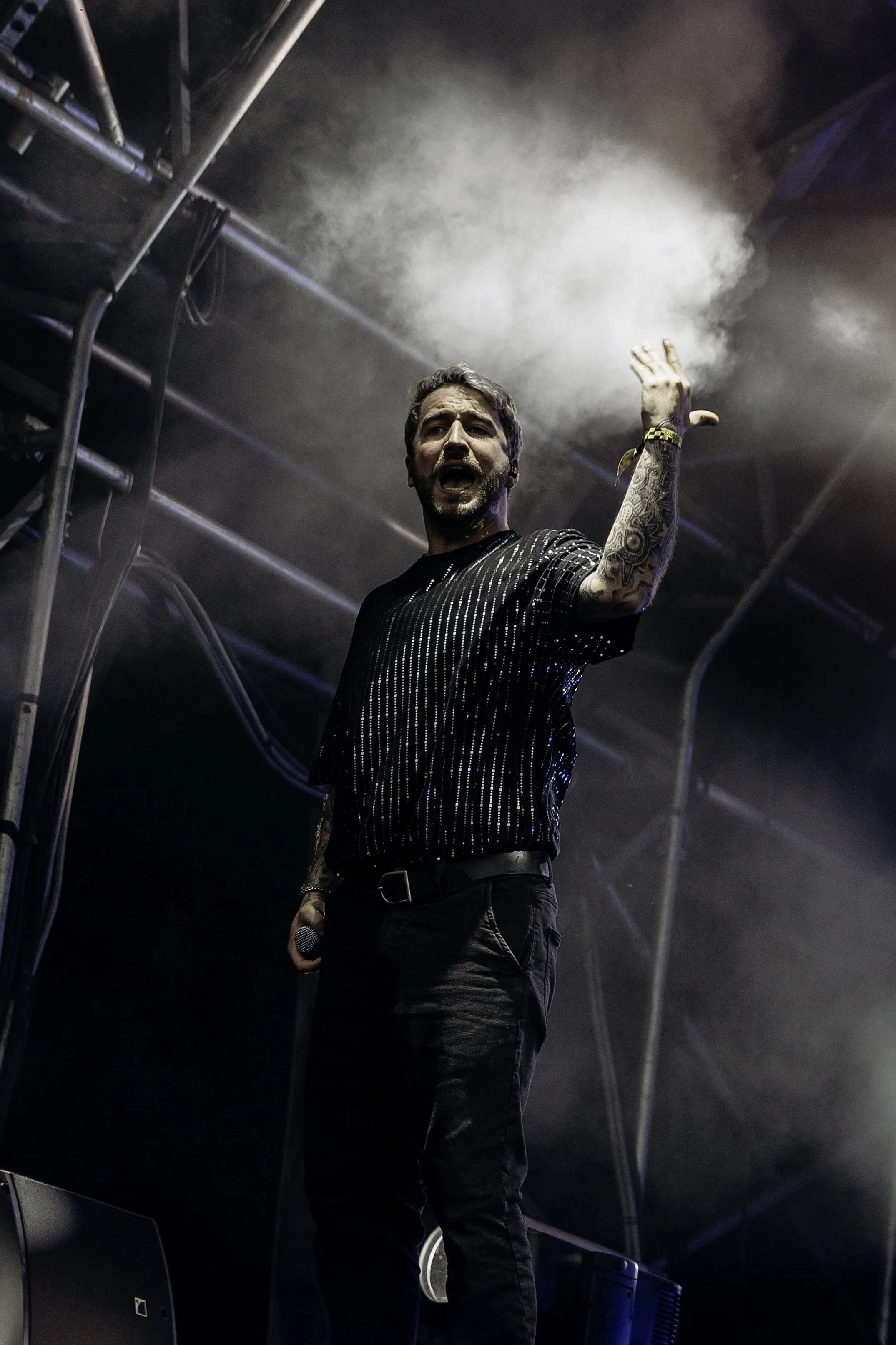 Male singer performing on stage with microphone, wearing a black shirt with white stripes, tattoos visible on his arms, and gesturing with his right hand.