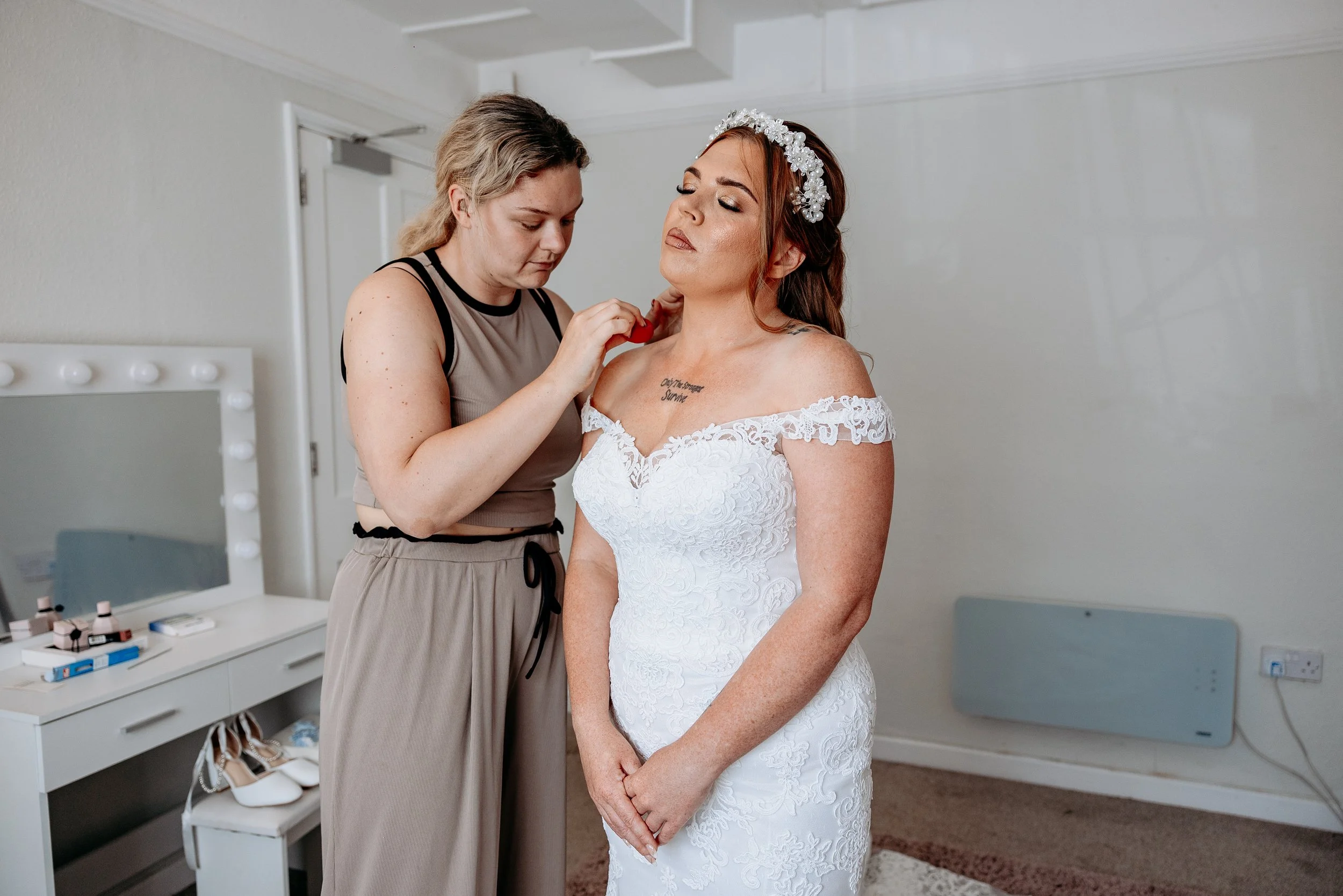 A bride with red hair in a white lace wedding dress and a floral headband has makeup applied by a woman in a beige dress in a room with a white makeup vanity and shoes on a stool.