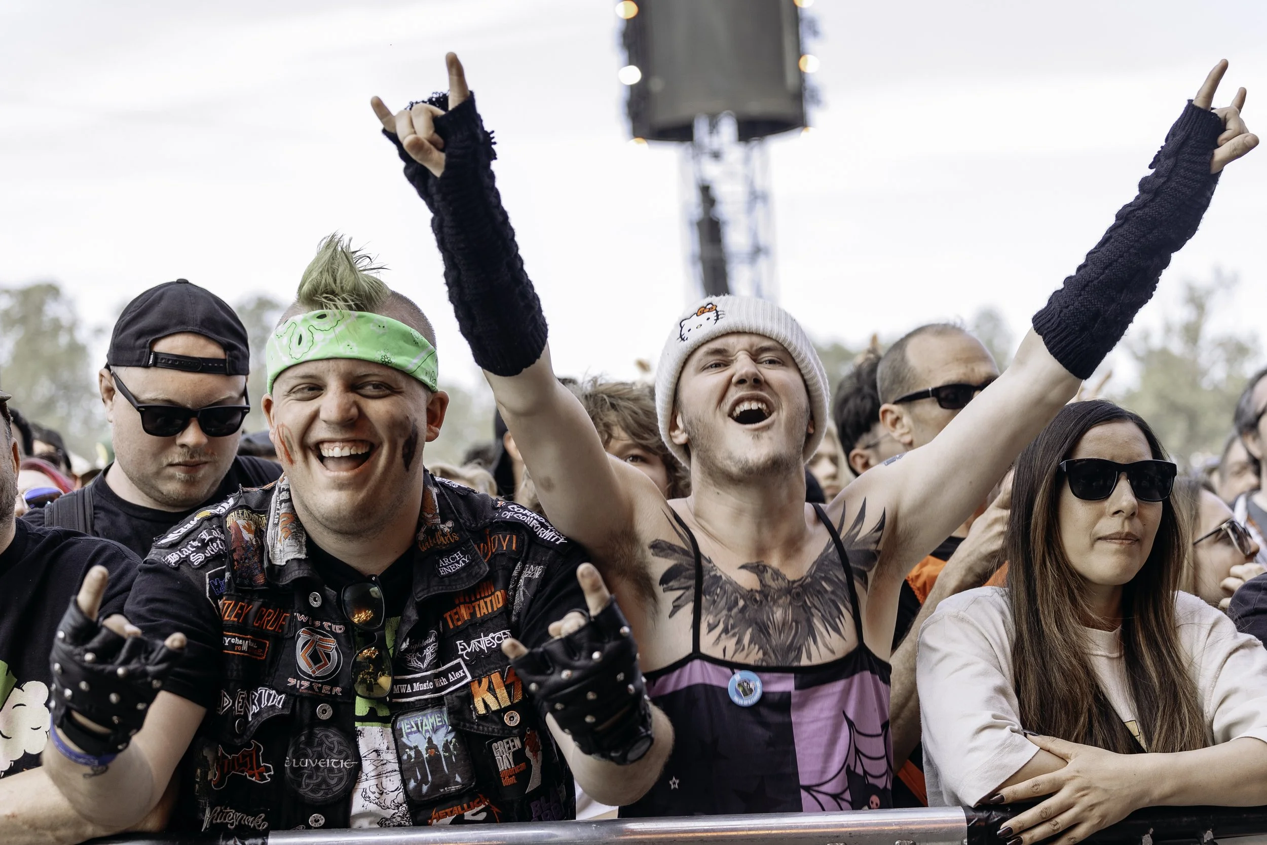 Crowd of people at a concert or festival, some smiling and making hand gestures, with a person in the center wearing a white knit hat with a Hello Kitty logo and showing enthusiasm.