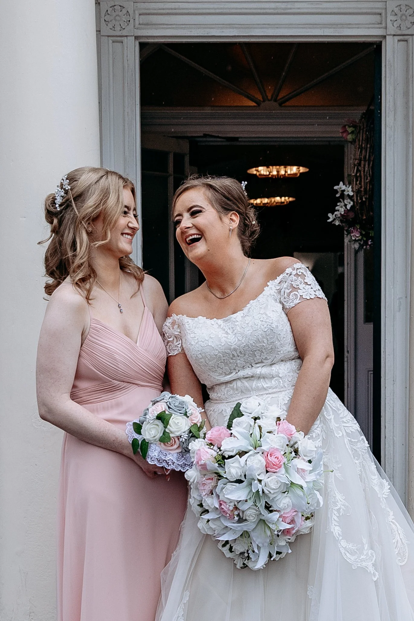 Two women, one in a white wedding dress and the other in a pink bridesmaid dress, smiling and sharing a joyful moment at a wedding, holding floral bouquets.