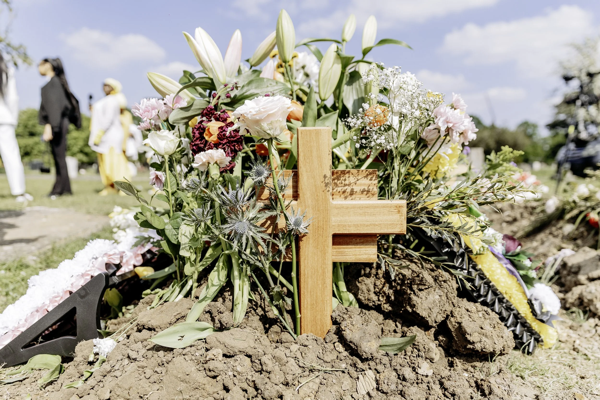 Flowers and a wooden cross on a mound of dirt at a grave site, with people in the background.