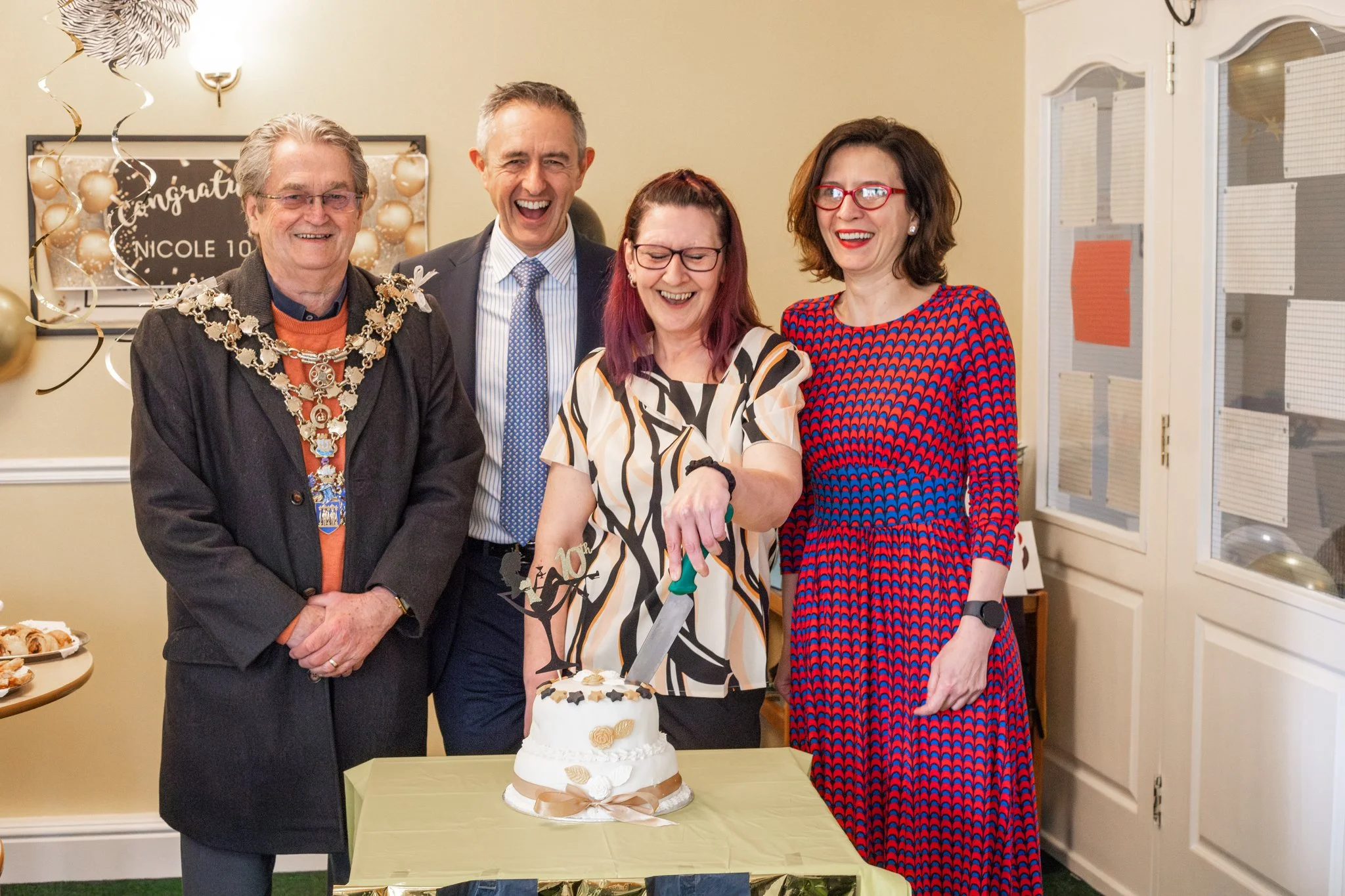 Group of four people celebrating a birthday with a cake, one woman cutting the cake with a knife, decorated with balloons and a banner in the background.