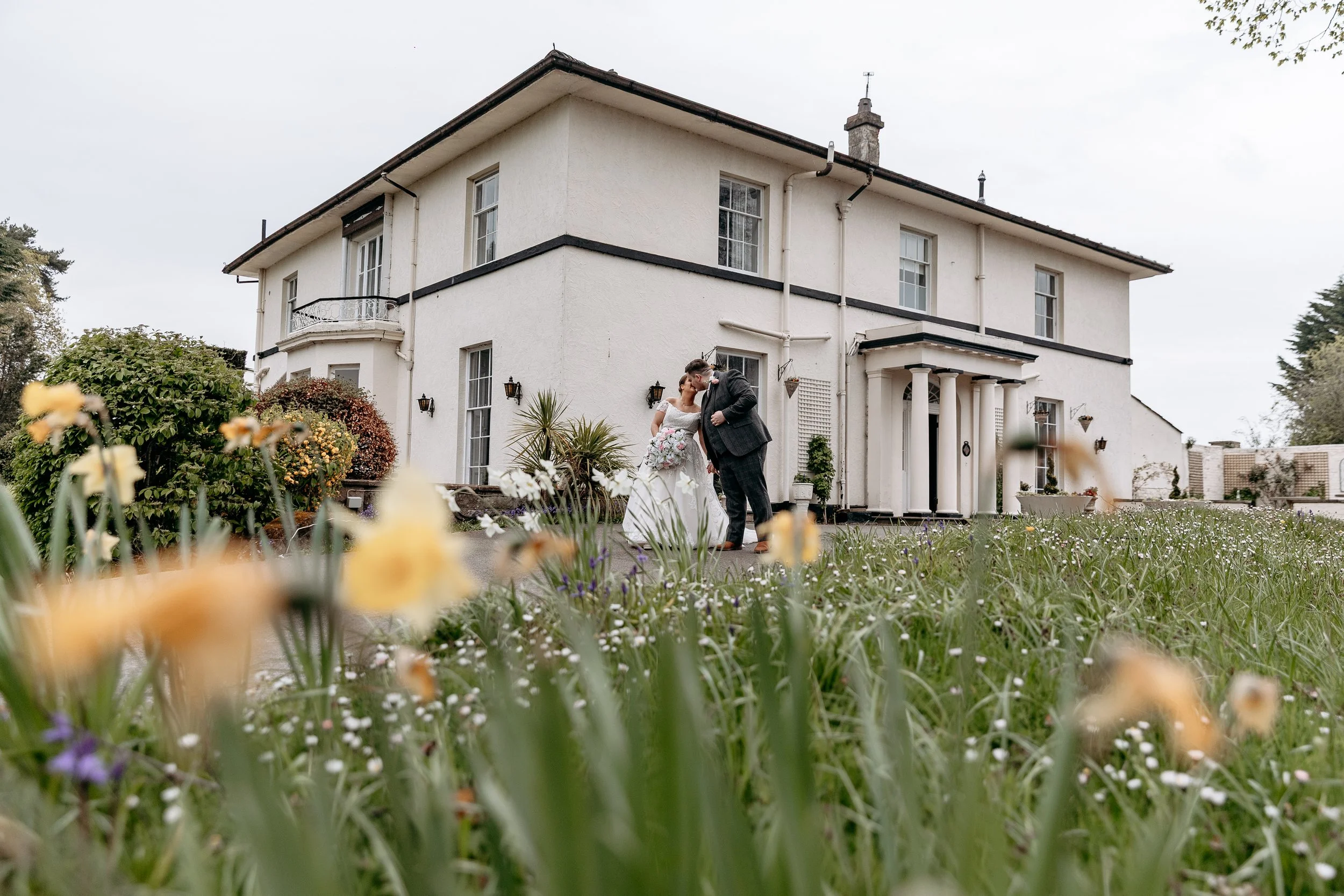 Bride and groom sharing a kiss outside a large white house surrounded by a garden with flowers.
