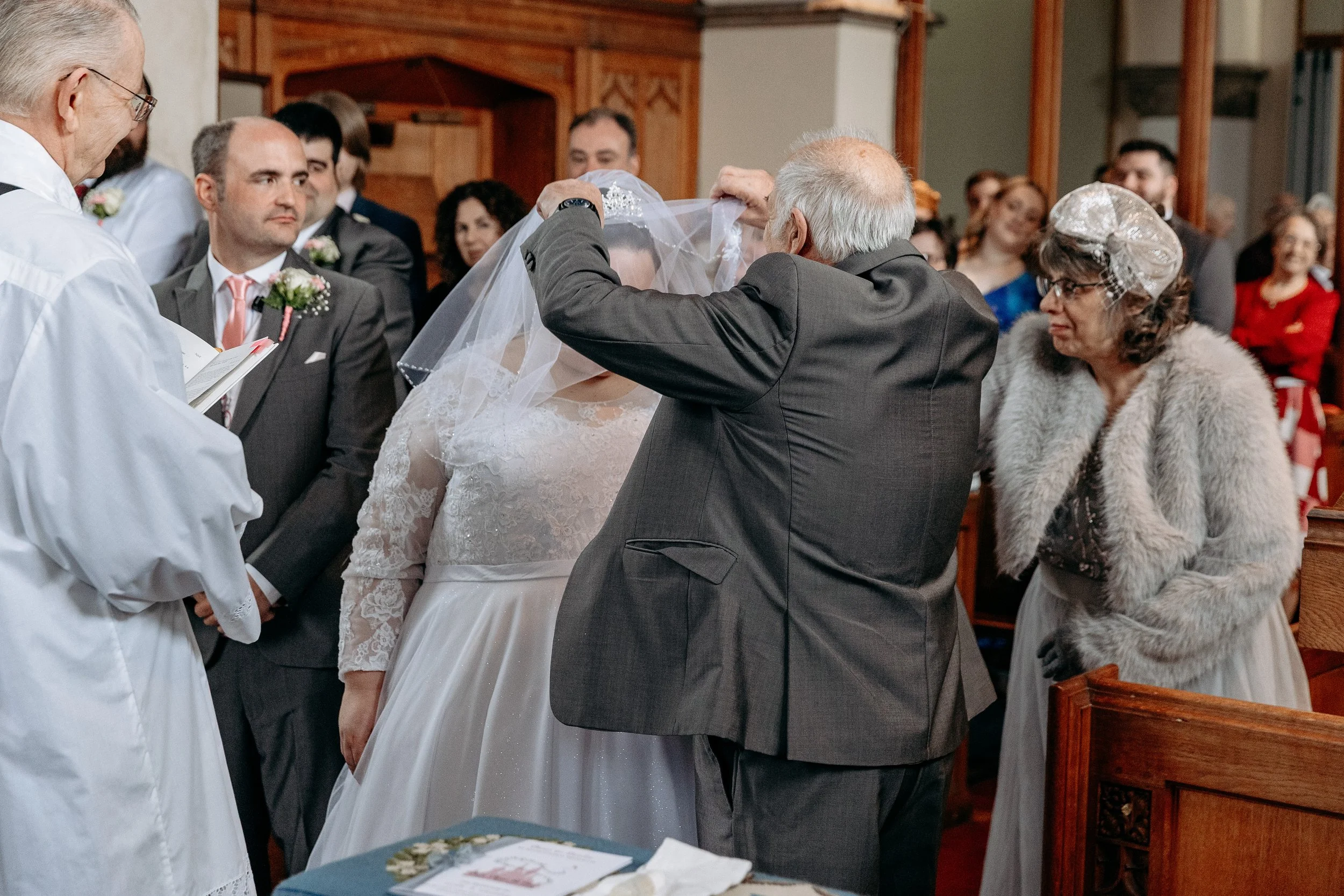 A wedding ceremony inside a church where an elderly man is lifting a bride's veil while a woman in a fur coat and headpiece looks on. Several guests watch in the background.