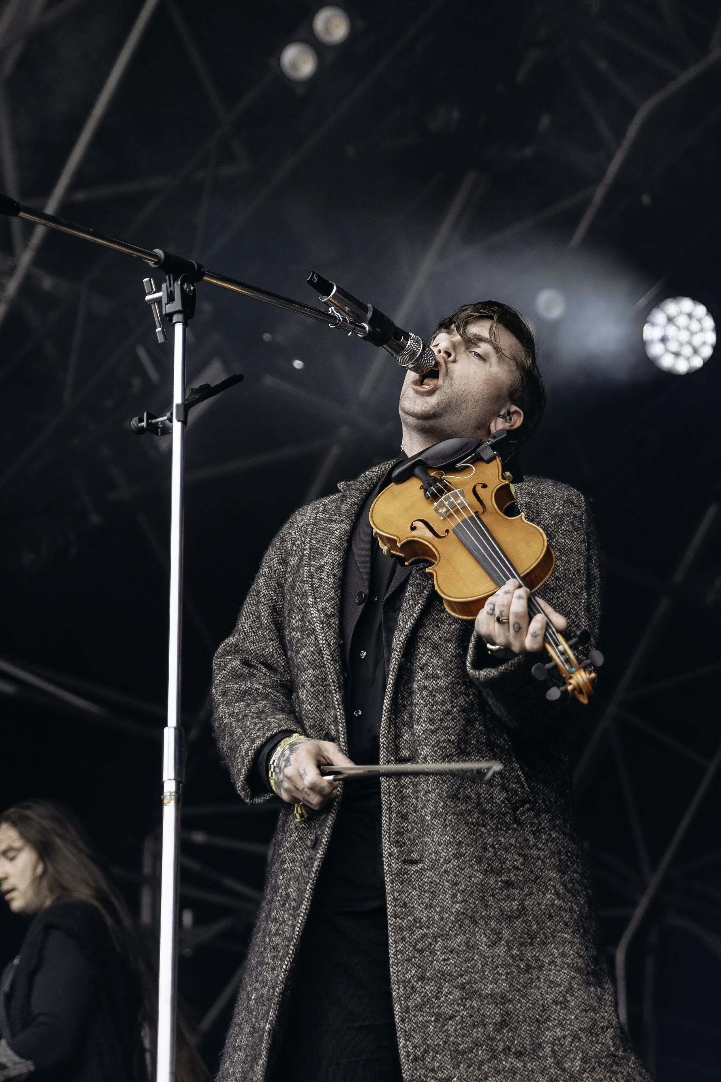 A male musician singing into a microphone and playing a violin on a stage, with another person in the background.