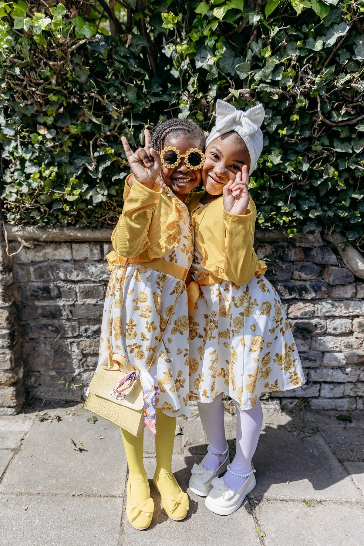 Two young girls smiling and dressed in matching yellow and white floral dresses, standing outdoors in front of a brick wall and green leafy background. One girl is wearing yellow tights and shoes with bows, sunglasses, and carrying a yellow purse, wh