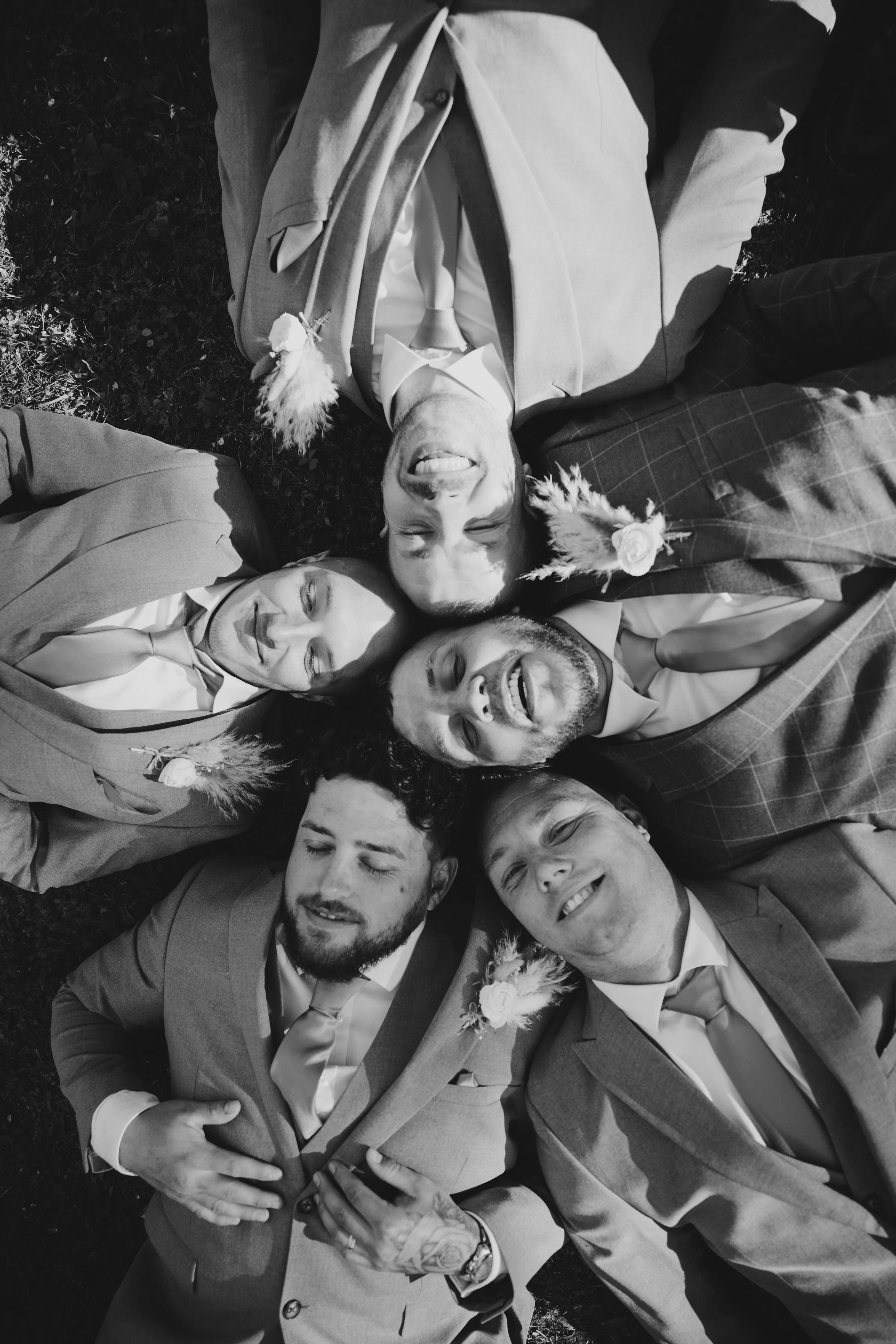 Black and white photo of five smiling people lying on the grass with their heads together, dressed in formal suits with boutonnières, showing joy and camaraderie.
