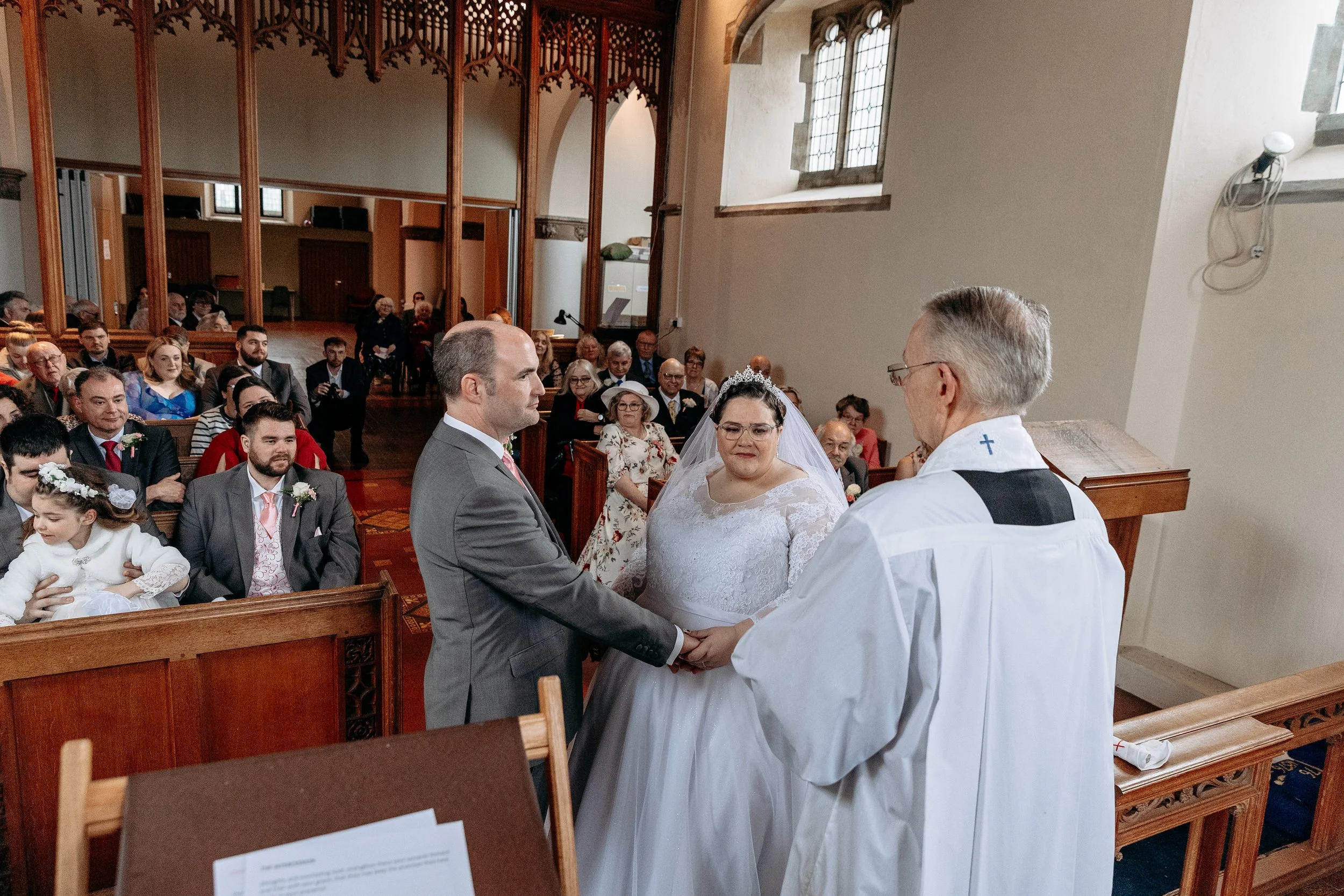 Wedding ceremony inside a church with a couple holding hands and facing a priest, with guests seated in pews.