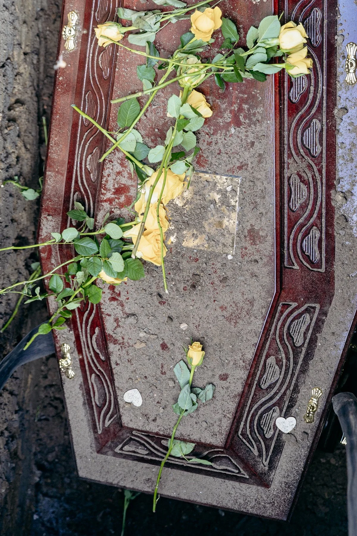 A gravesite with a decorative stone border adorned with yellow roses laid across the surface. A single yellow rose lies separately at the foot of the grave, surrounded by small heart-shaped tokens. The soil is visible around the grave.
