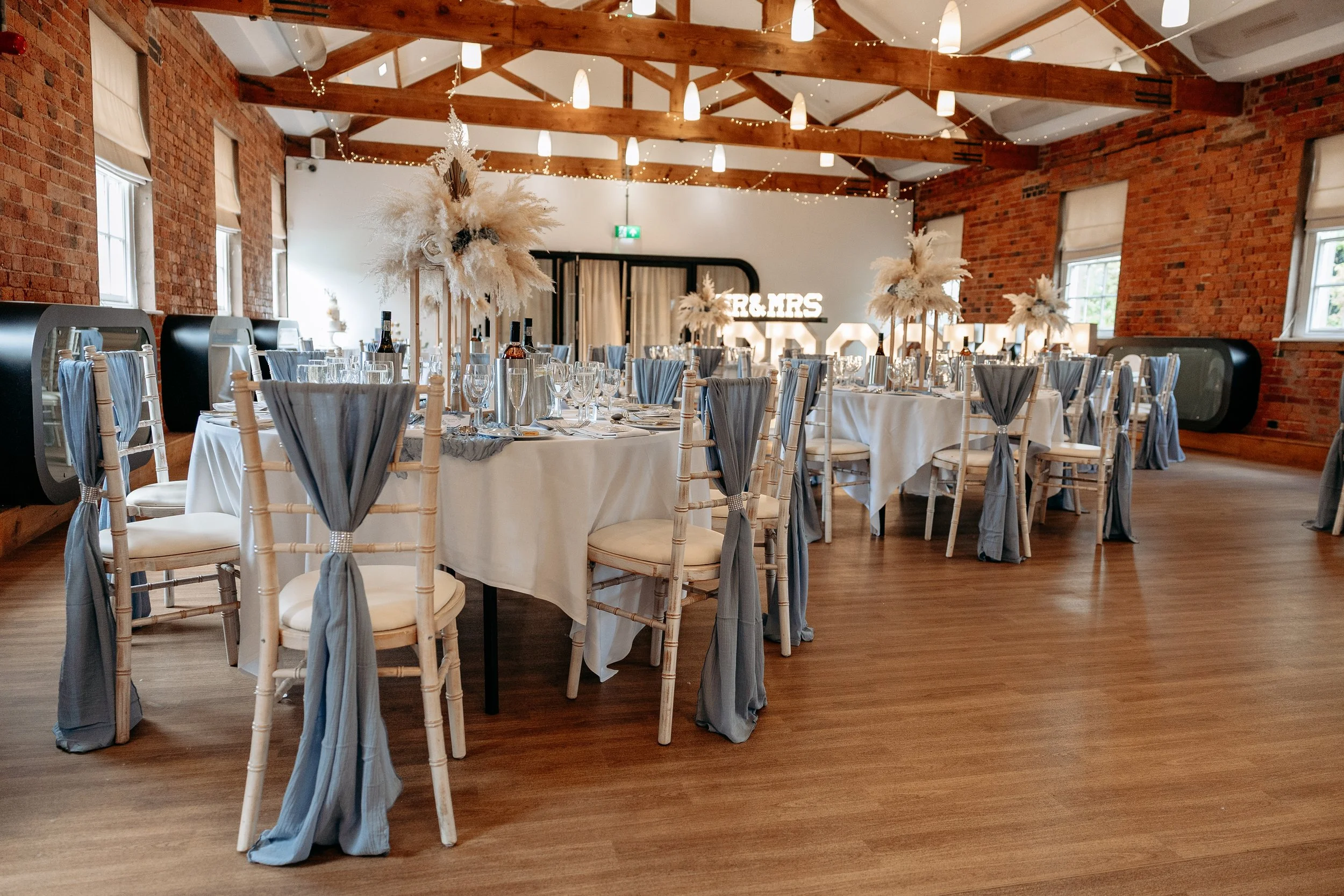 Decorated wedding reception in a rustic venue with round tables, white tablecloths, tall floral centerpieces, and chairs with gray drapes, featuring exposed brick walls and wooden beams.