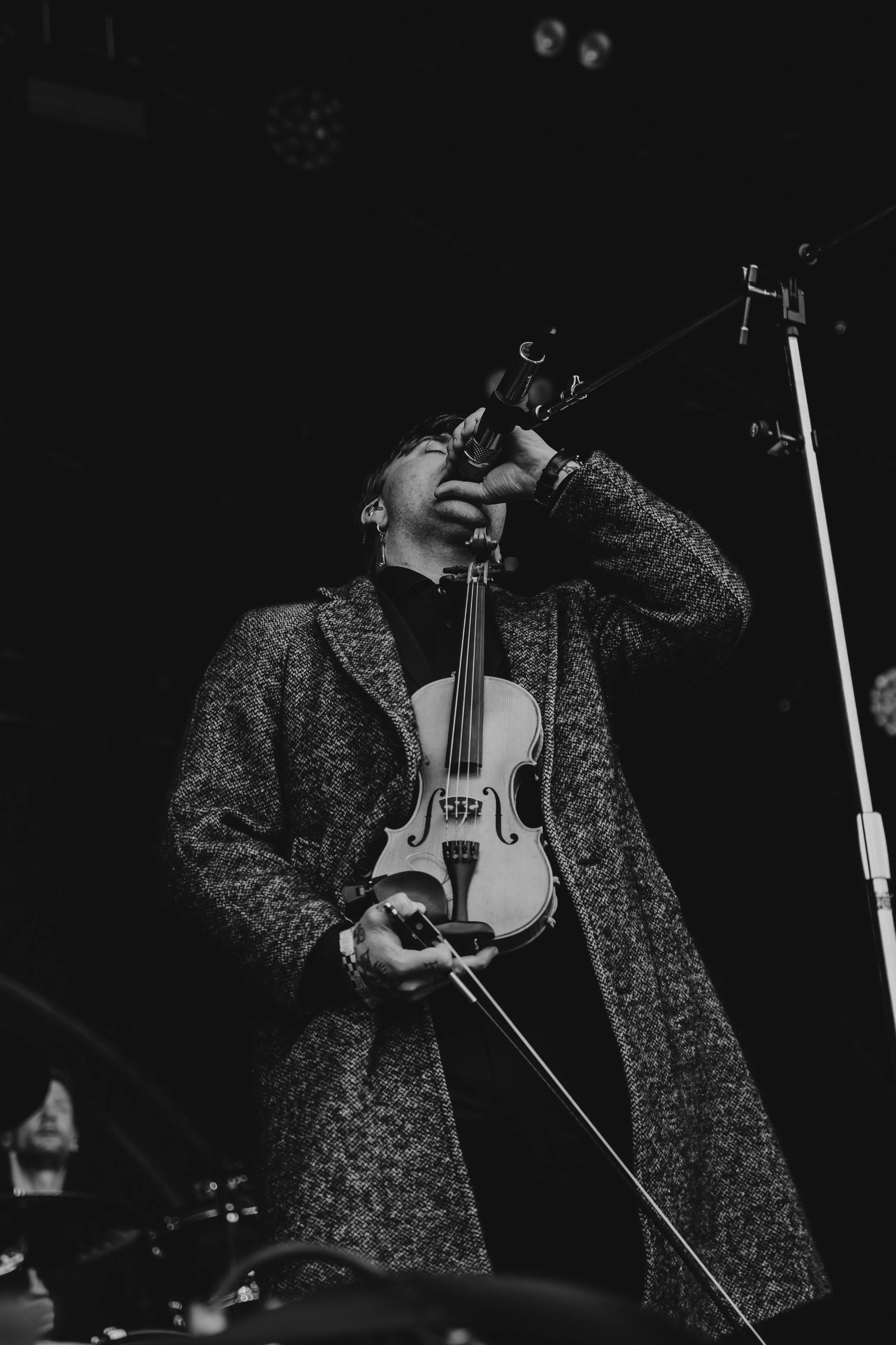 Black and white photo of a male musician performing on stage, holding a microphone with one hand and a violin with the other, wearing a long coat.