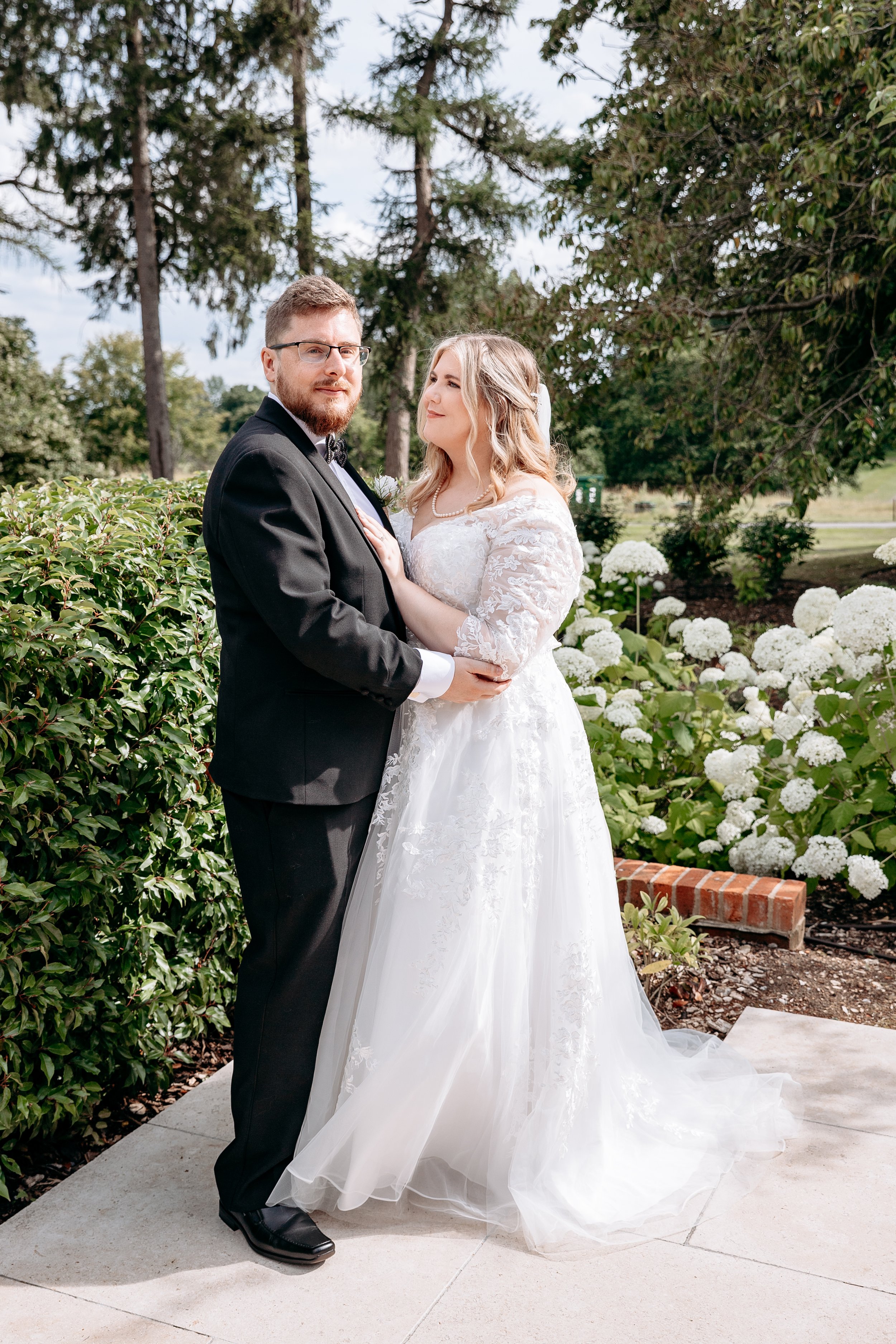 Bride in white lace dress and groom in black tuxedo embracing in a garden setting with hydrangeas and trees.