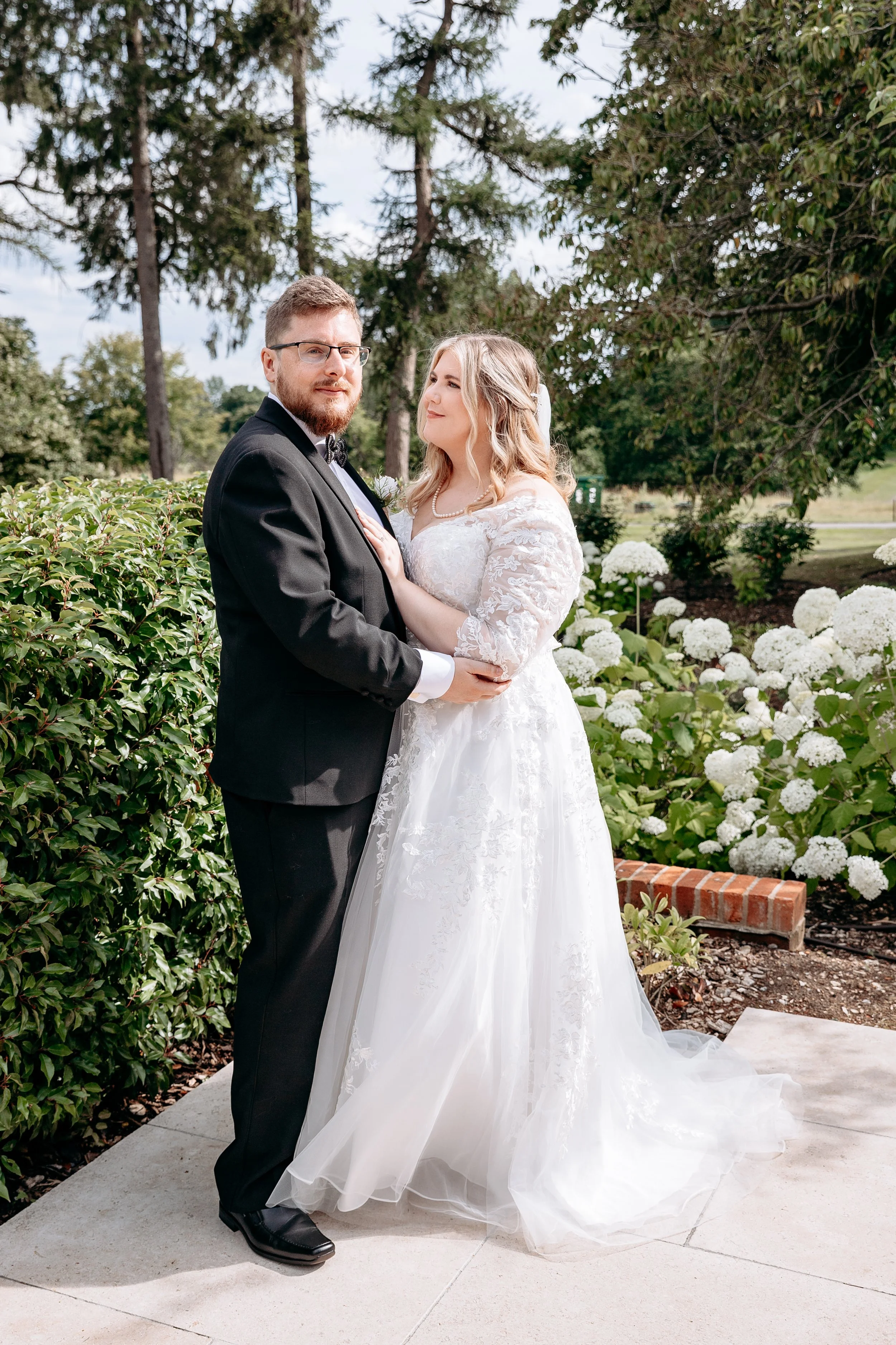 Bride in white lace dress and groom in black tuxedo embracing in a garden setting with hydrangeas and trees.