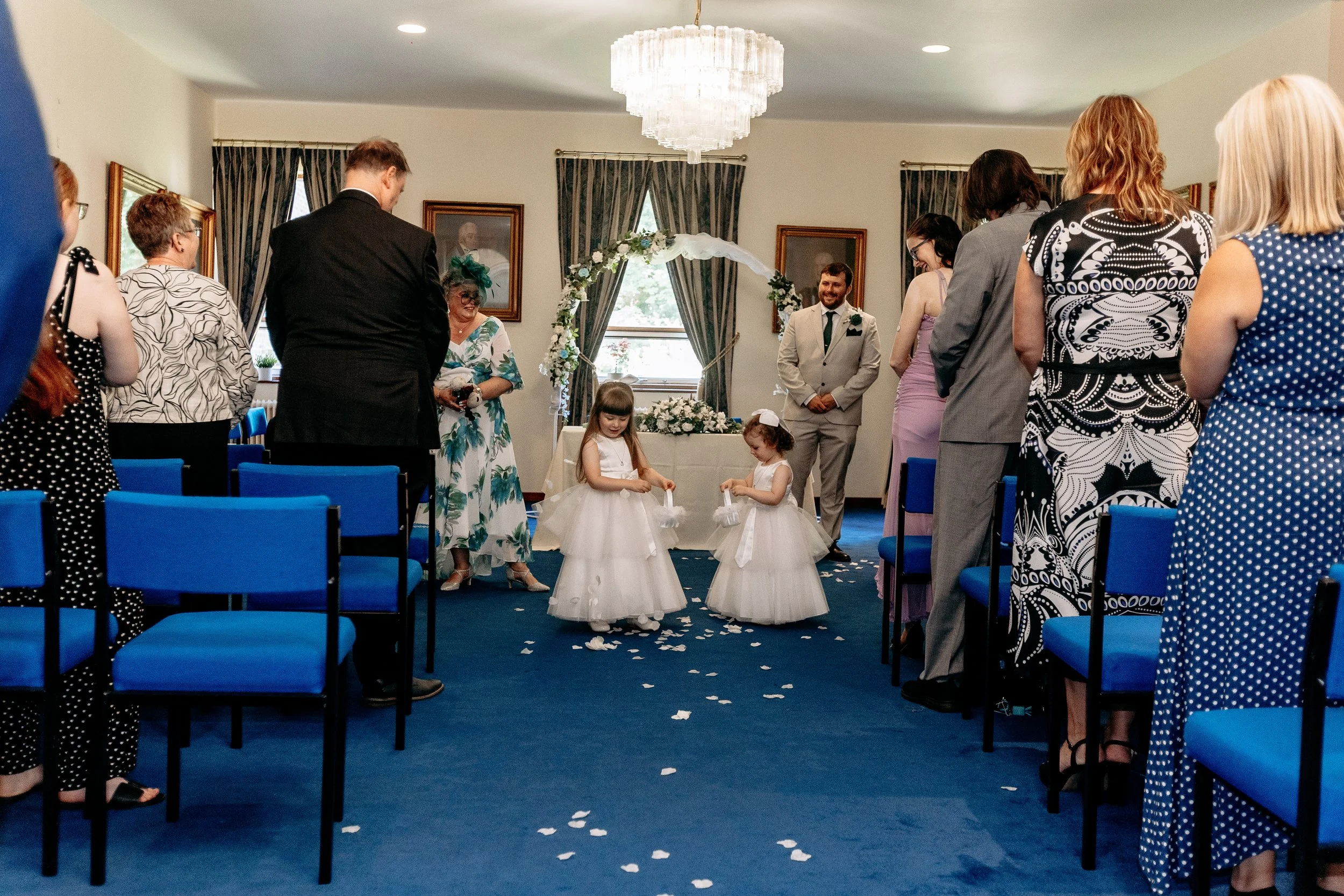 A wedding ceremony inside a decorated room with guests standing, two young flower girls in white dresses scattering flower petals, and the bride and groom smiling at each other.