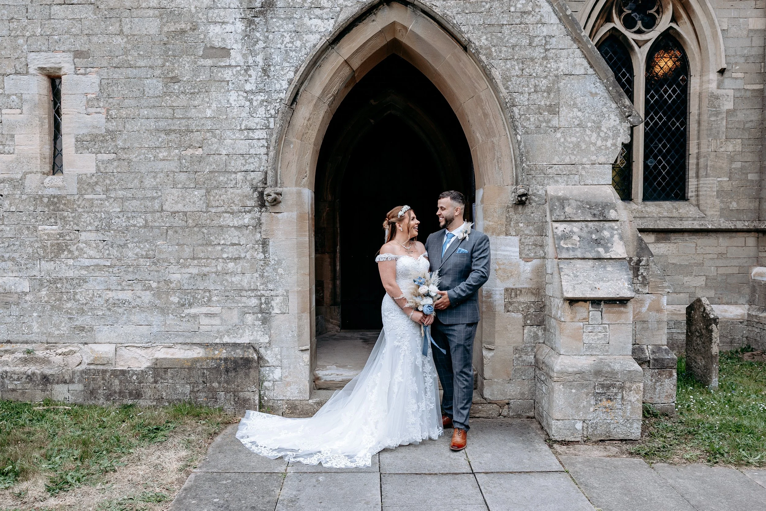 Bride and groom standing outside a stone church, smiling at each other, with the bride holding a bouquet and wearing a white wedding gown, and the groom in a gray suit with a blue tie.