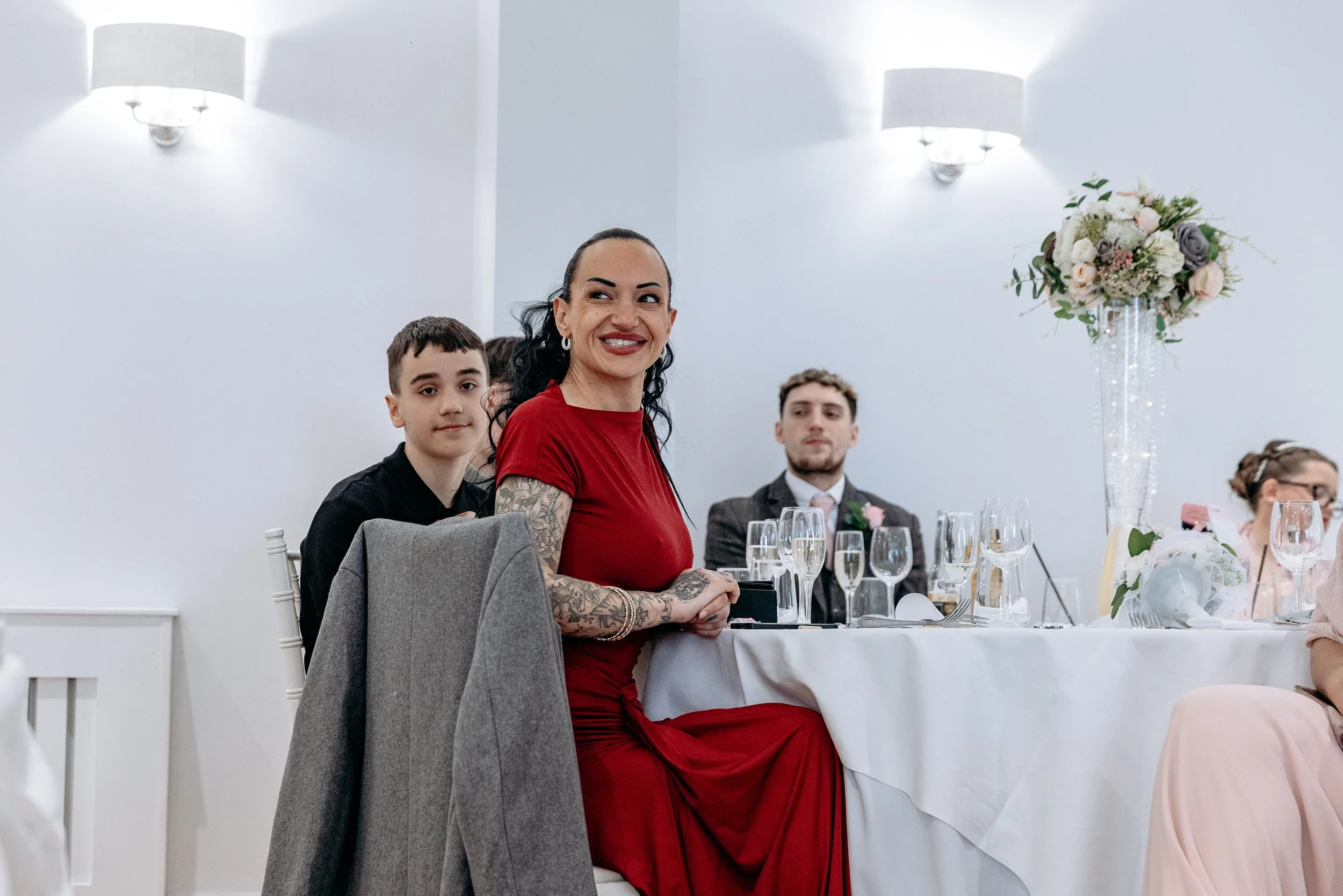 A woman in a red dress smiling at a formal event, seated at a table with two young men and decorated with flowers and wine glasses.