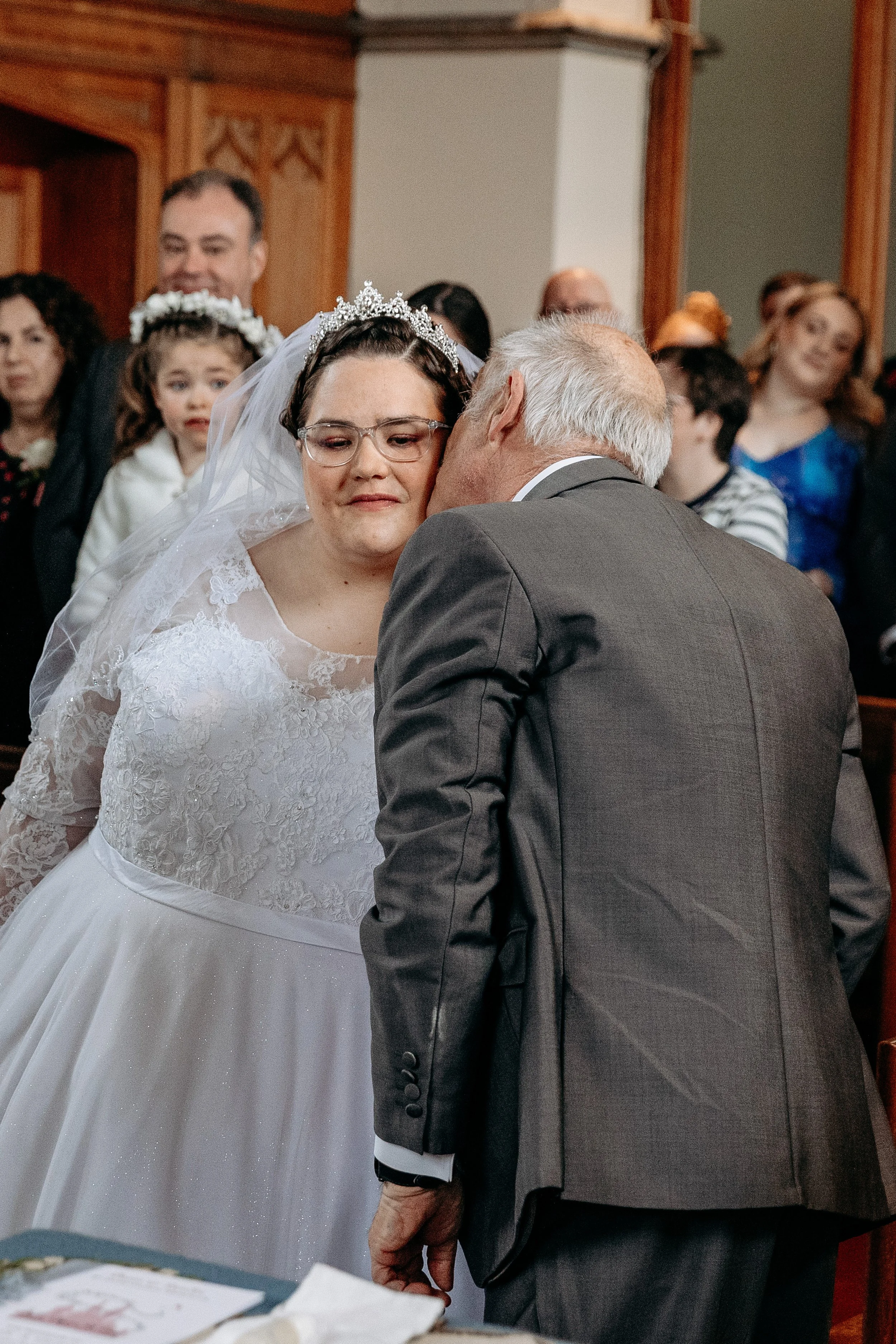 A bride with glasses and a tiara, wearing a white lace wedding dress, is being kissed on the cheek by an older man in a gray suit at a wedding ceremony, with guests watching in the background.