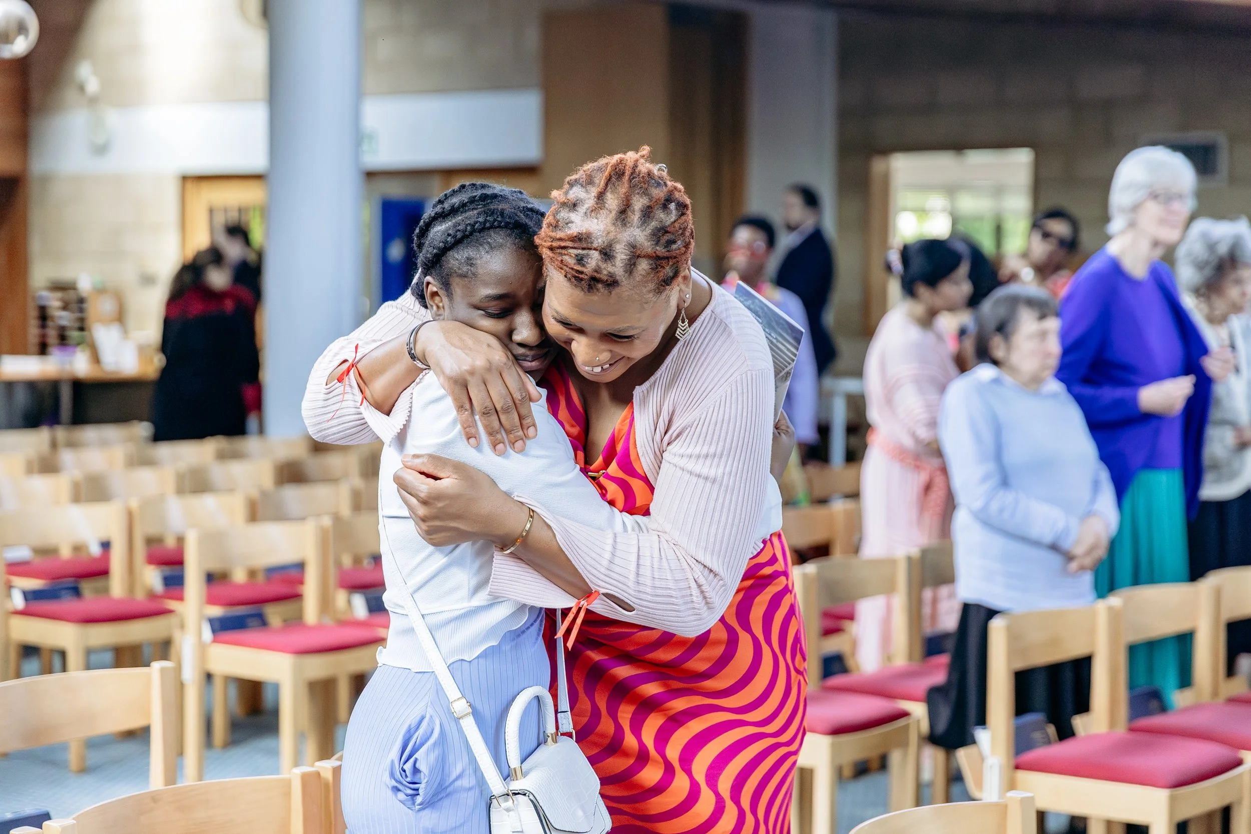 Two women hugging each other warmly in a church or gathering space, with chairs and other people in the background.
