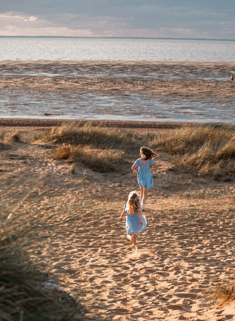Two children in blue dresses running on a sandy beach with the ocean in the background under a cloudy sky.