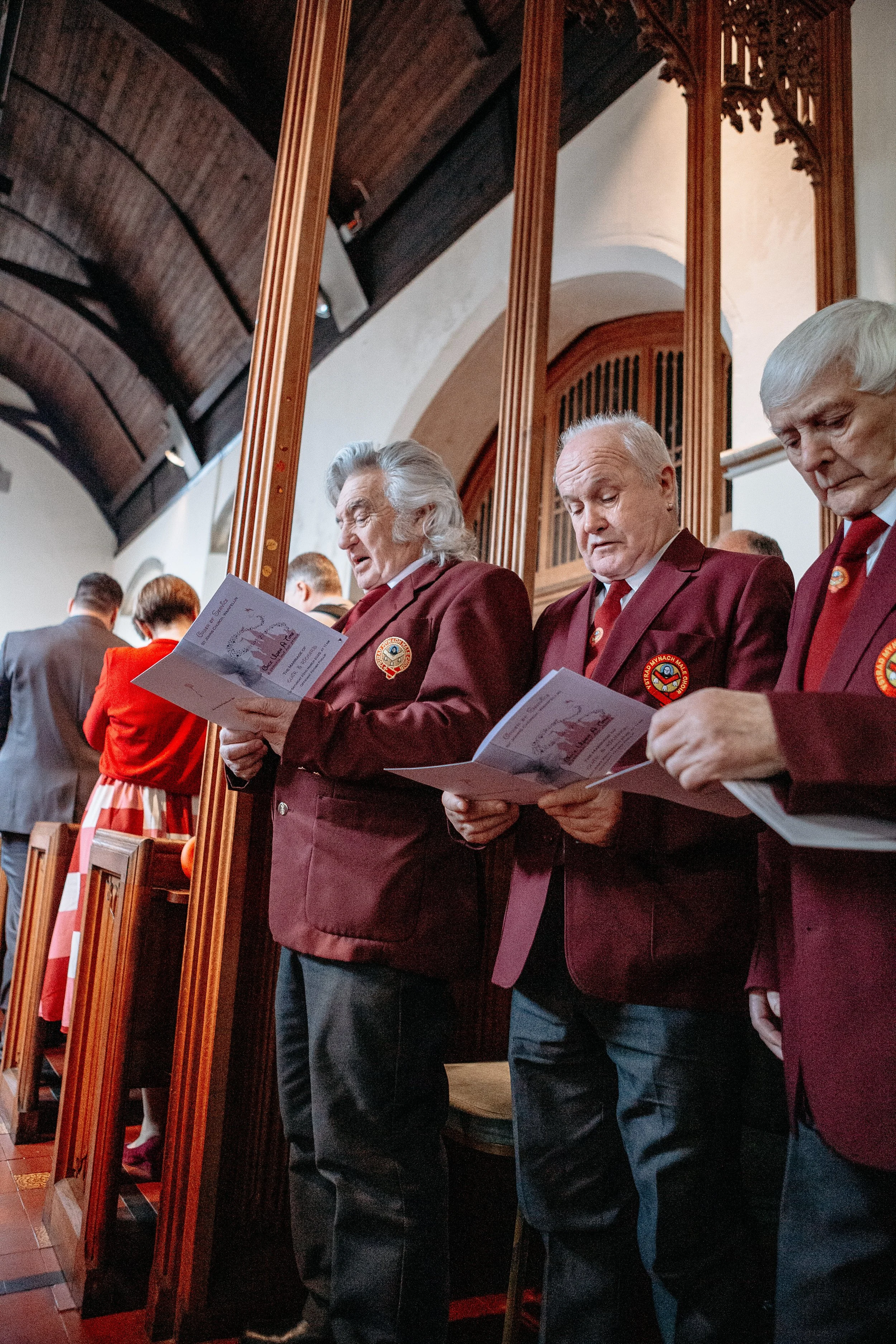 Three elderly men in maroon blazers and red ties standing in a church, holding and reading programs during a service or ceremony.