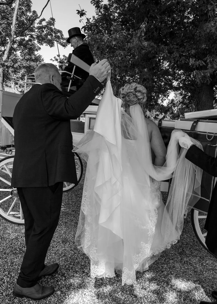 A bride dressed in a wedding gown and veil is getting into a horse-drawn carriage with the help of two men, one holding her dress and the other holding a flag, while a man in a top hat is sitting inside the carriage. Trees are visible in the backgrou
