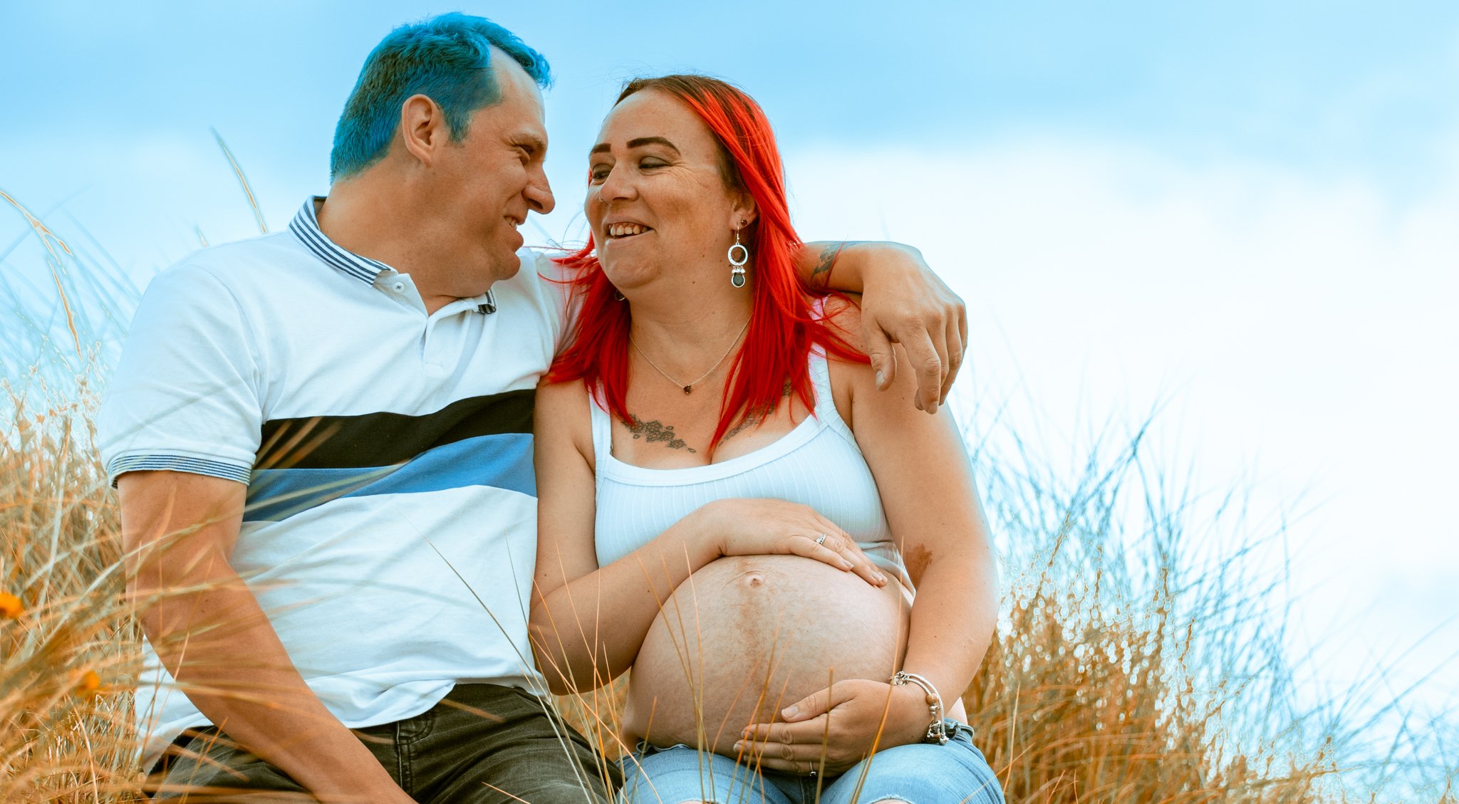 A pregnant woman with bright red hair and a man with short blue hair sitting in a field of tall, dry grass, smiling at each other, the woman resting her hand on her belly.