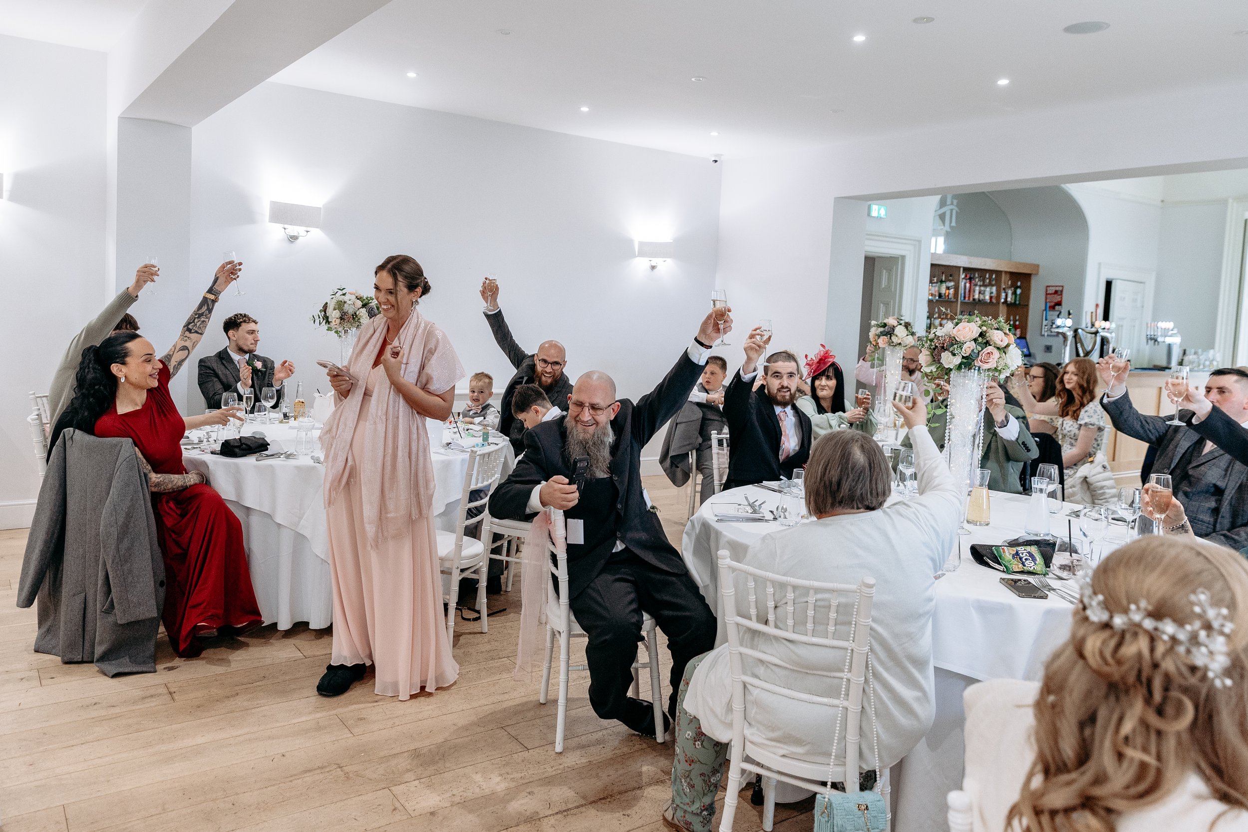 People celebrating at a wedding reception, raising glasses in toast around decorated tables with floral centerpieces in a white-walled banquet hall.