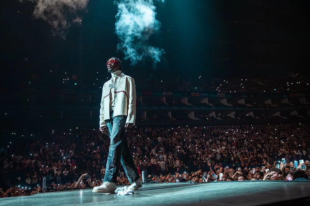 Performer with white jacket, blue patterned pants, and red glasses on stage with smoke, large audience in a crowded indoor concert venue.