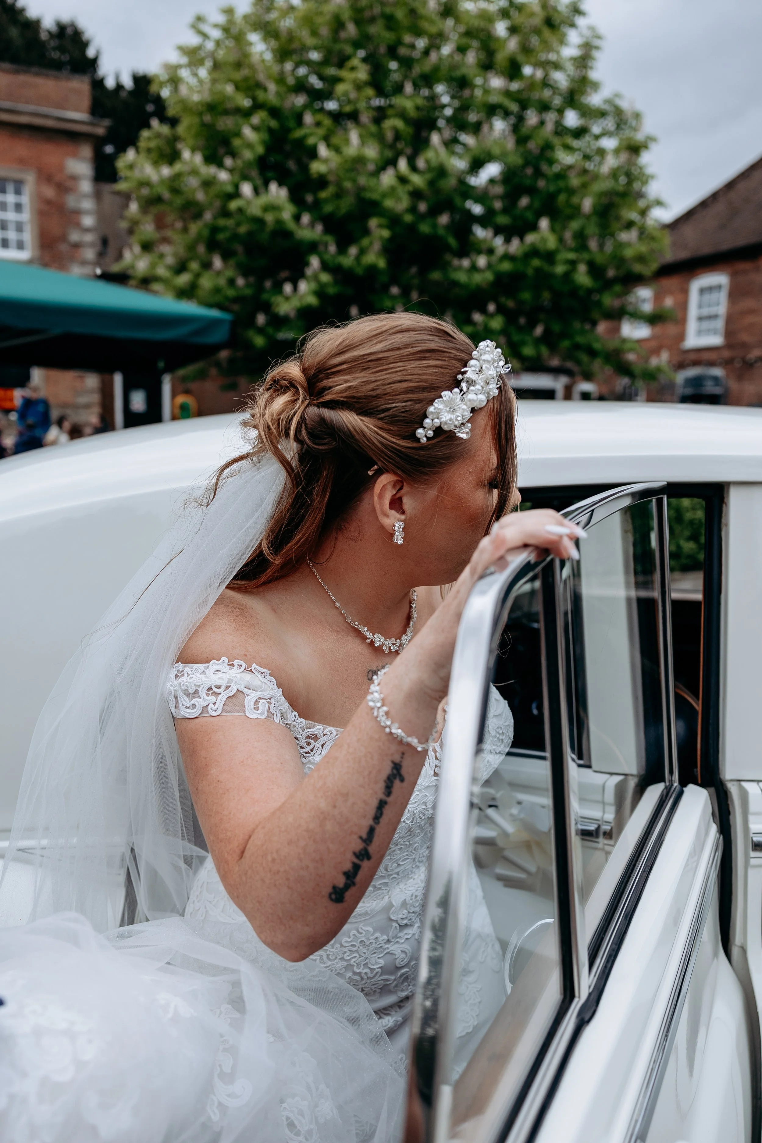 Bride in a white wedding dress with lace details, wearing pearl jewelry, and a floral headpiece, getting into a white vintage car outside with trees and brick buildings in the background.