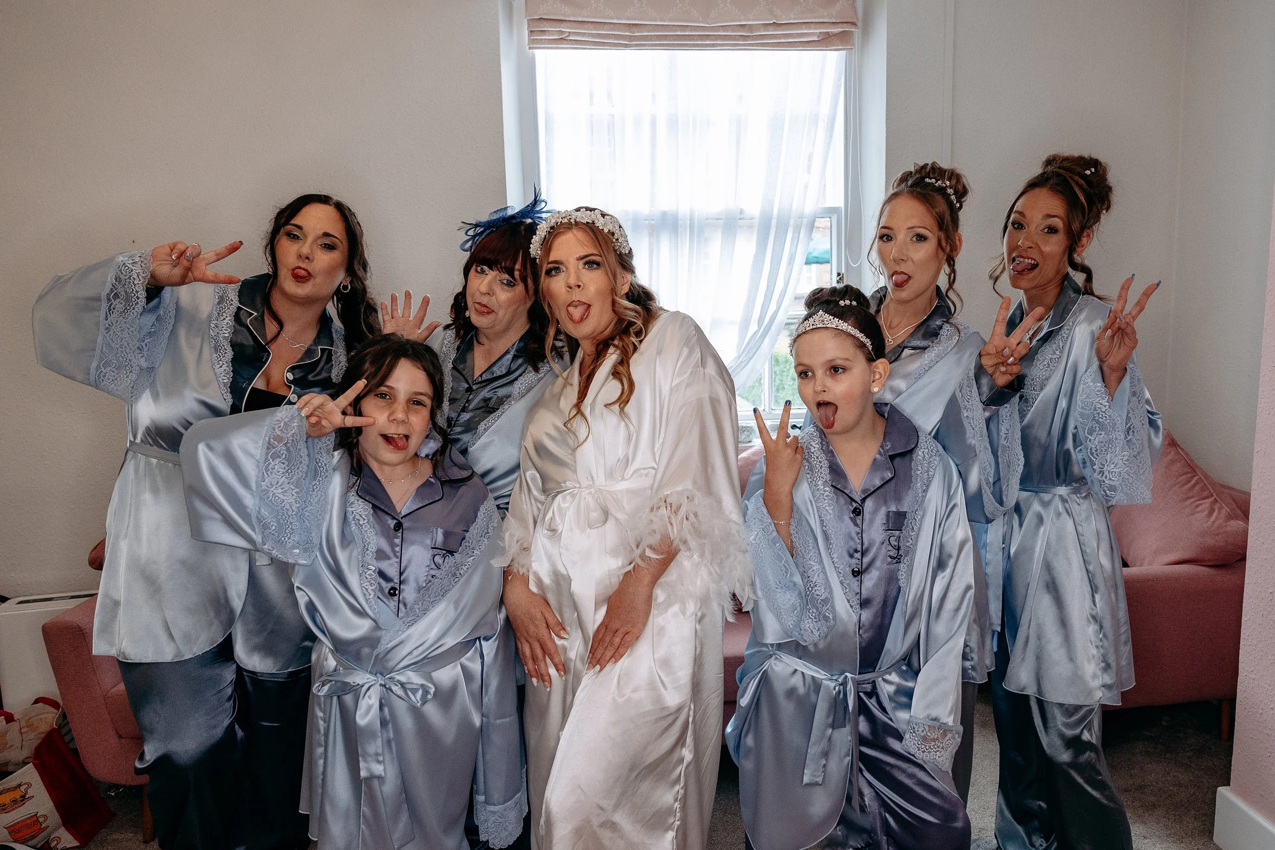 A group of women and girls in matching satin pajamas making silly faces and peace signs in a room with a pink sofa and window in the background.