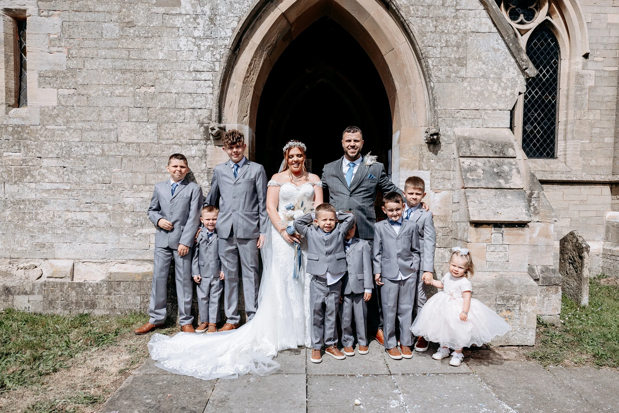 A wedding party standing on steps in front of a stone church. The bride and groom are in the center, with children around them, some smiling and posing for the photo.
