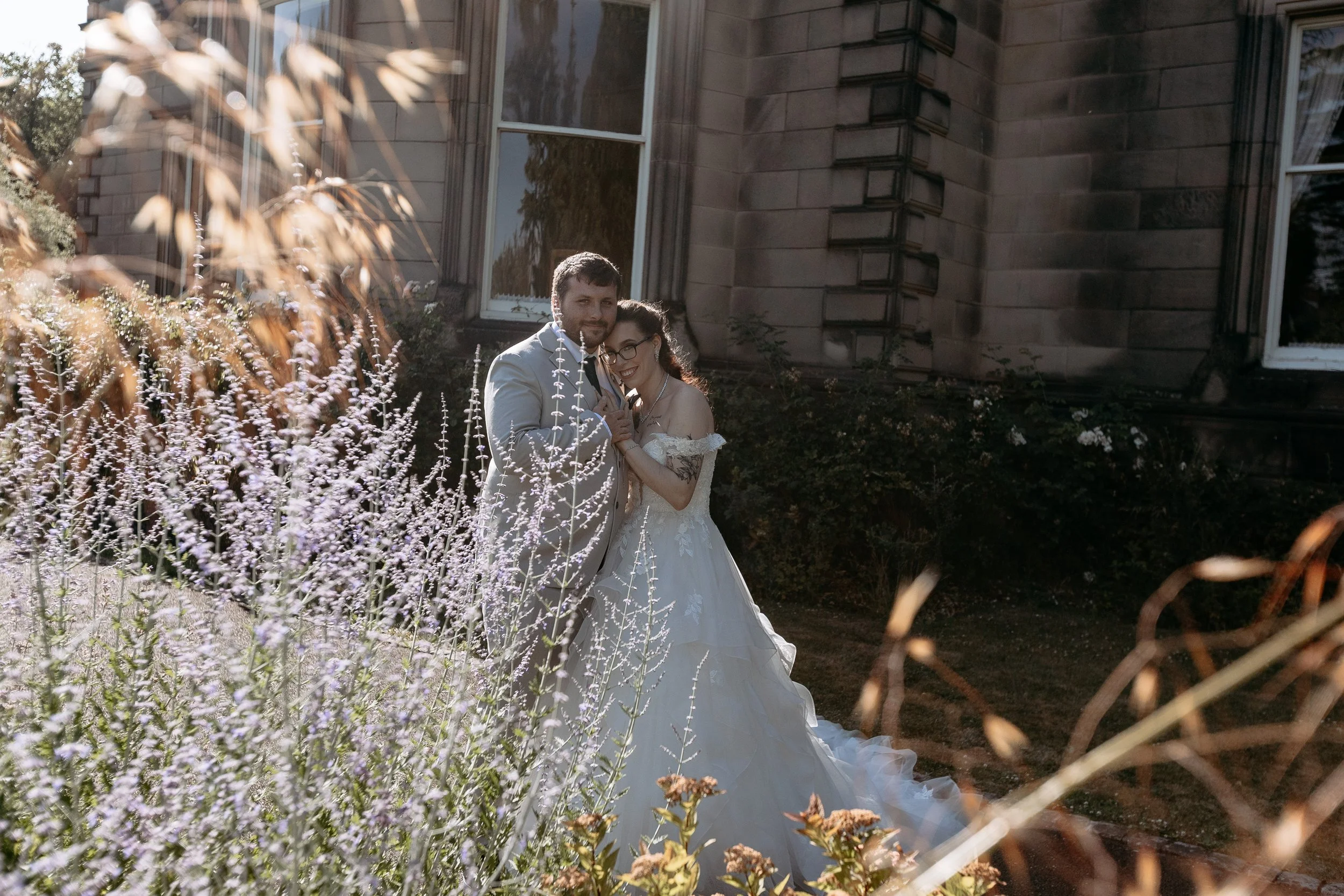 A bride and groom in wedding attire standing outside near flowering plants and a stone building, embracing and smiling.