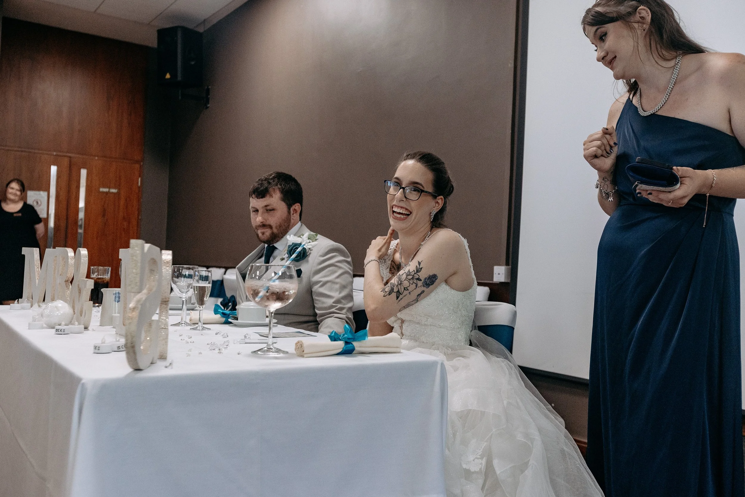 Bride in wedding dress with tattoos sitting at head table, smiling, next to groom in suit, with a woman in navy blue dress standing nearby during wedding reception.