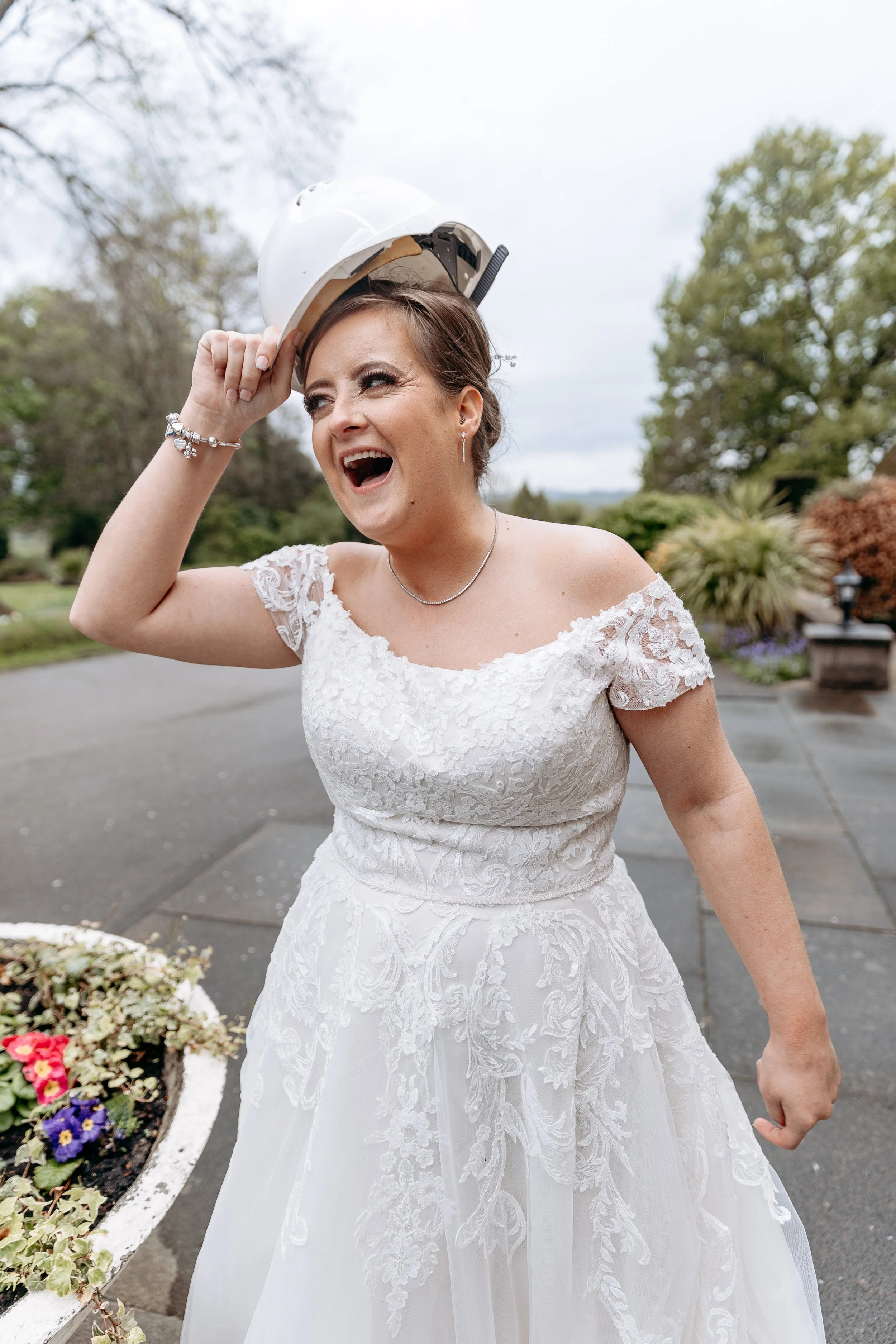 A woman in a wedding dress is smiling and putting on a white safety helmet outdoors.