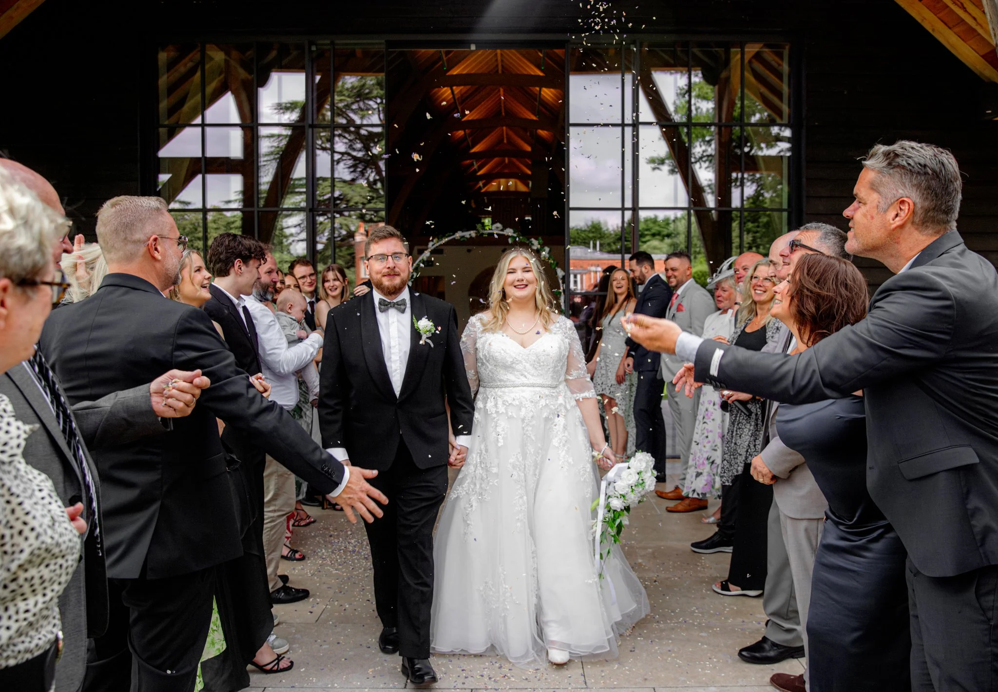 A bride and groom walking hand in hand through a crowd outside a building, surrounded by friends and family celebrating their wedding, with confetti in the air.