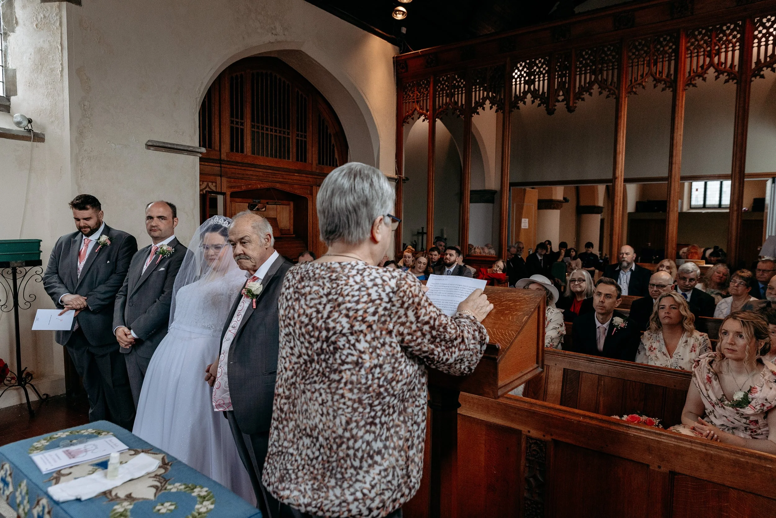 A bride and groom standing before a church officiant during their wedding ceremony, with family and friends seated in pews watching.