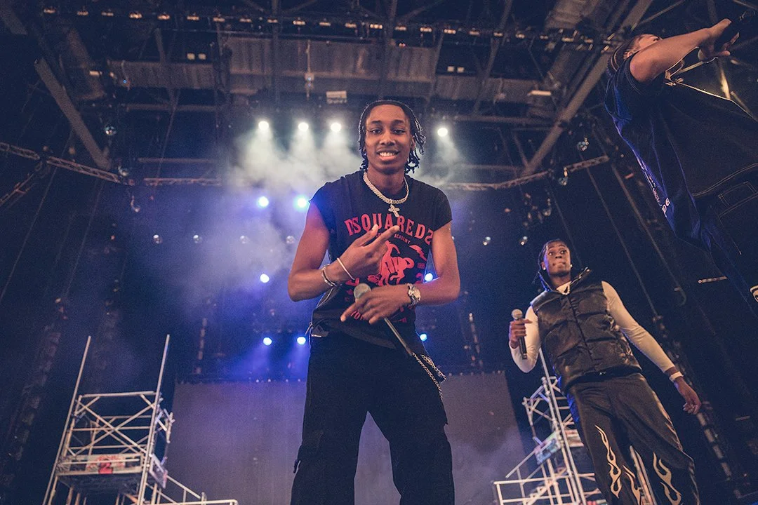 Three performers on stage with microphones, wearing casual outfits, under bright stage lights and surrounded by stage equipment.